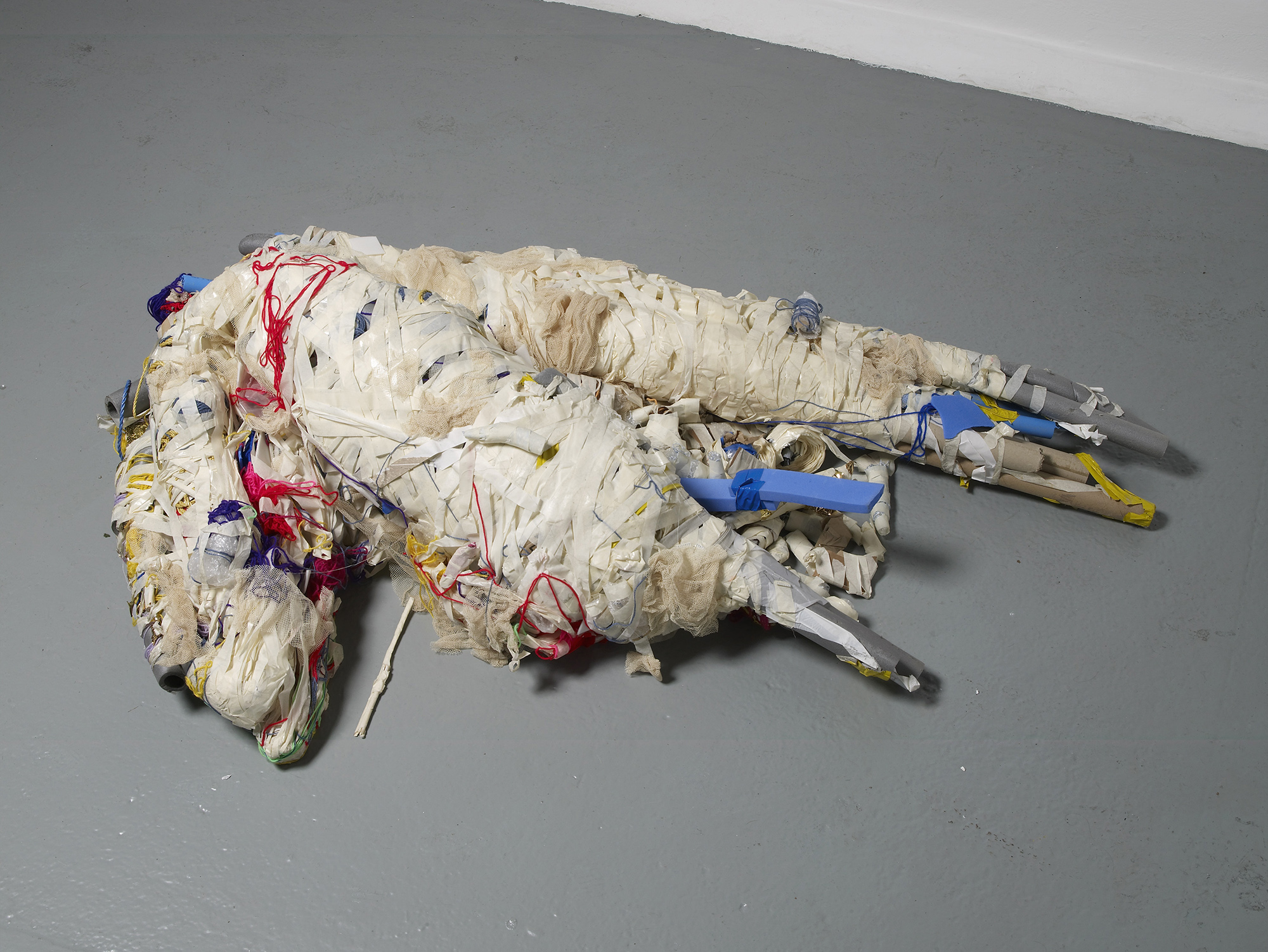 Two large, abstract forms made of white, wrapped fabric and plastic, with red, yellow, and blue threads and tubes, are lying intertwined on a grey floor in an art gallery setting.