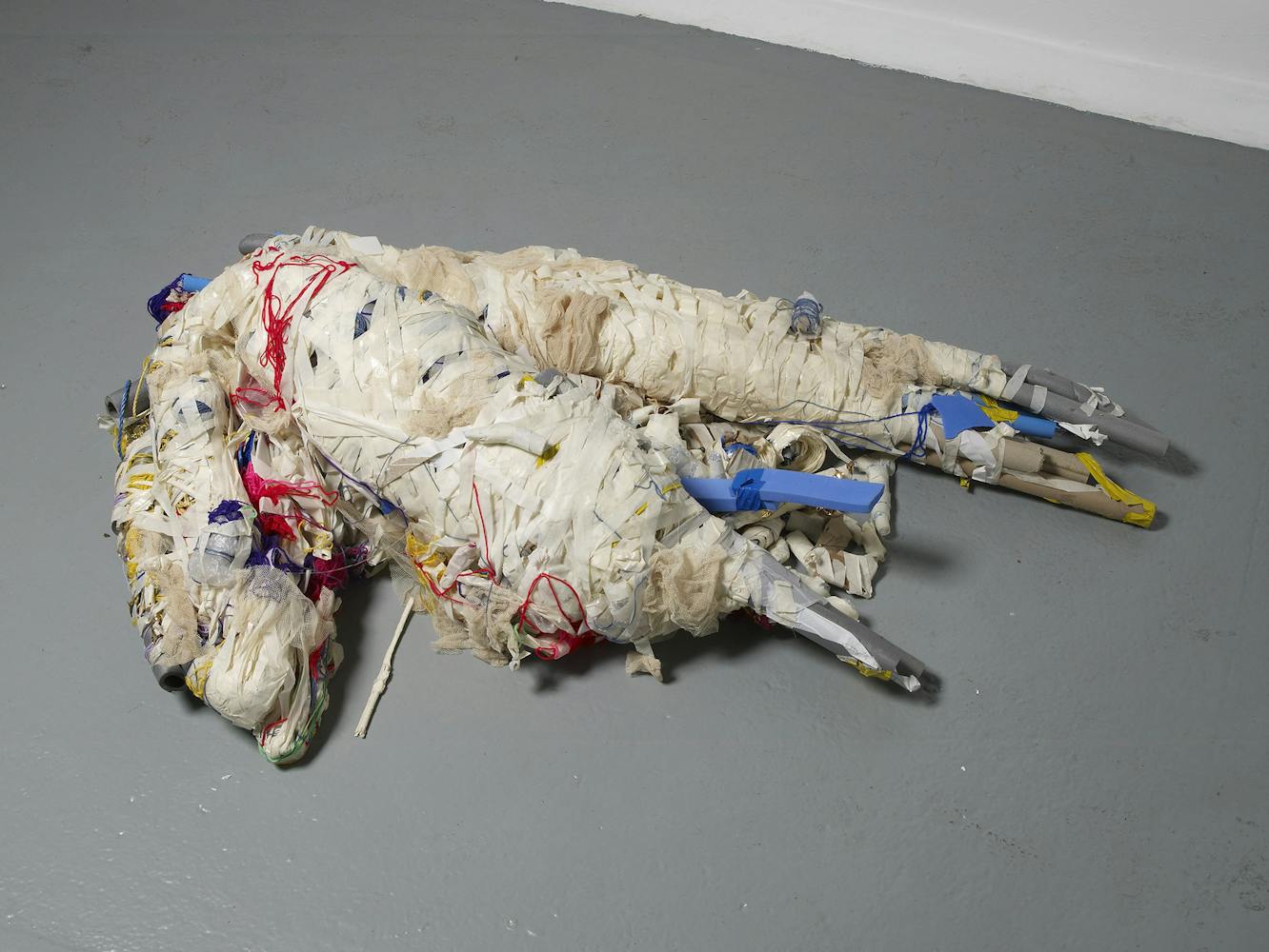 Two large, abstract forms made of white, wrapped fabric and plastic, with red, yellow, and blue threads and tubes, are lying intertwined on a grey floor in an art gallery setting.