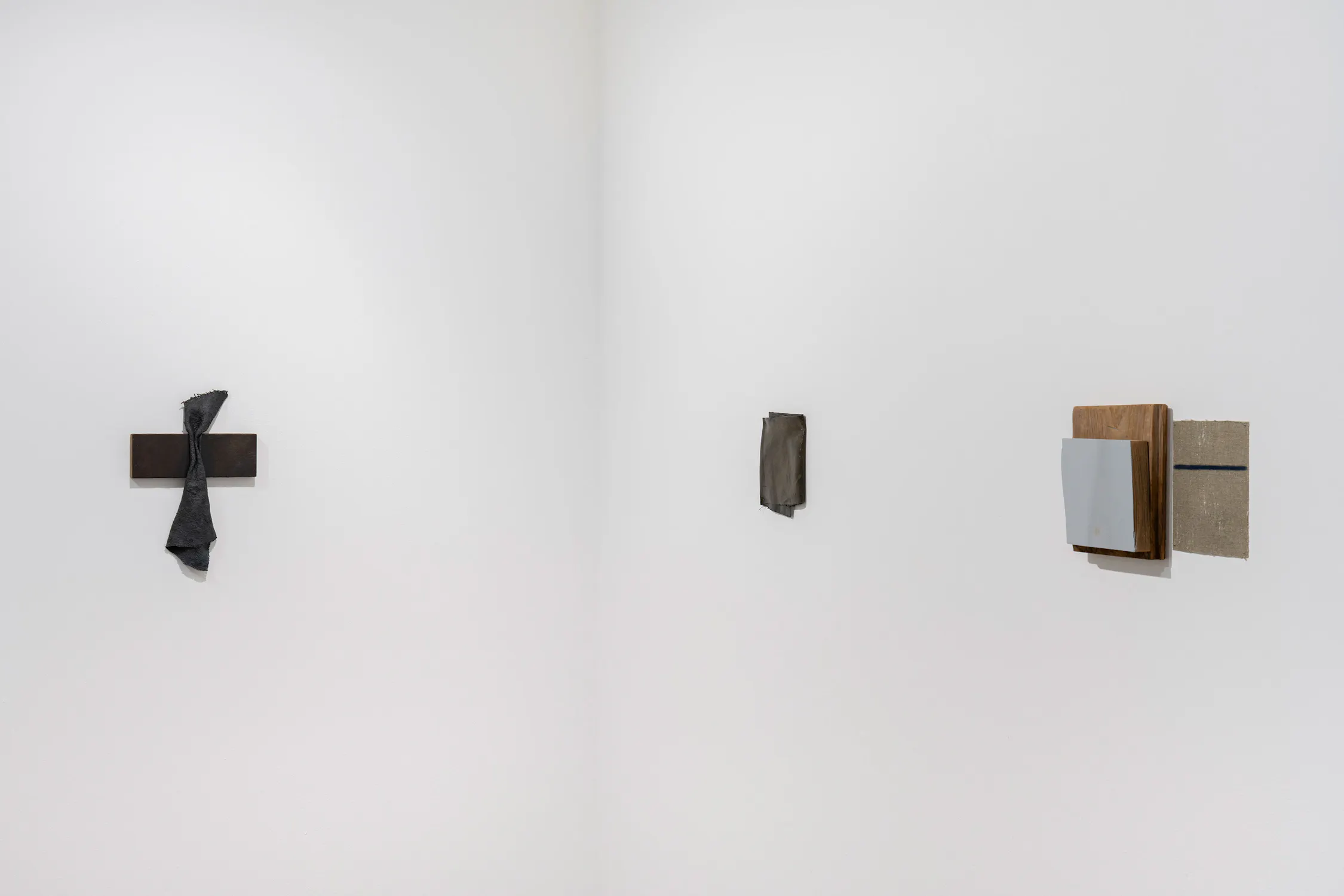 Three small abstract artworks by Bil Walton hang on a white corner wall. To the left is a dark cross draped with a grey cloth. In the centre is a small, rectangular grey panel. To the right is a layered composition featuring a light grey square, a dark wooden block, and a tan canvas.