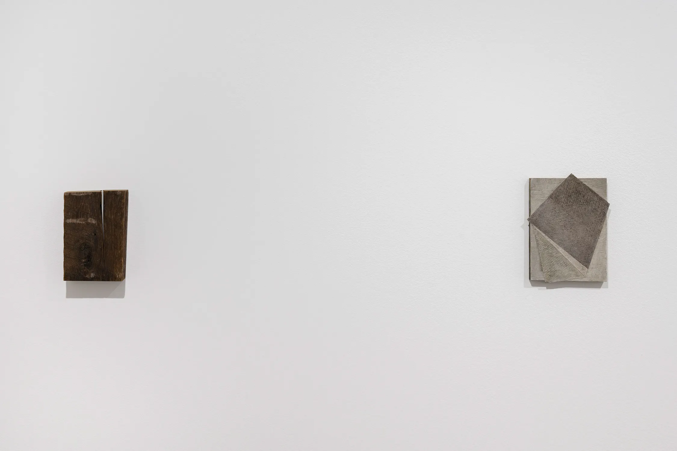 Two small, square-shaped artworks hang on a plain white wall. On the left is a thick, dark brown block of wood with a vertical grain. On the right is a flat, grey square featuring an abstract composition of overlapping geometric shapes with textured surfaces.