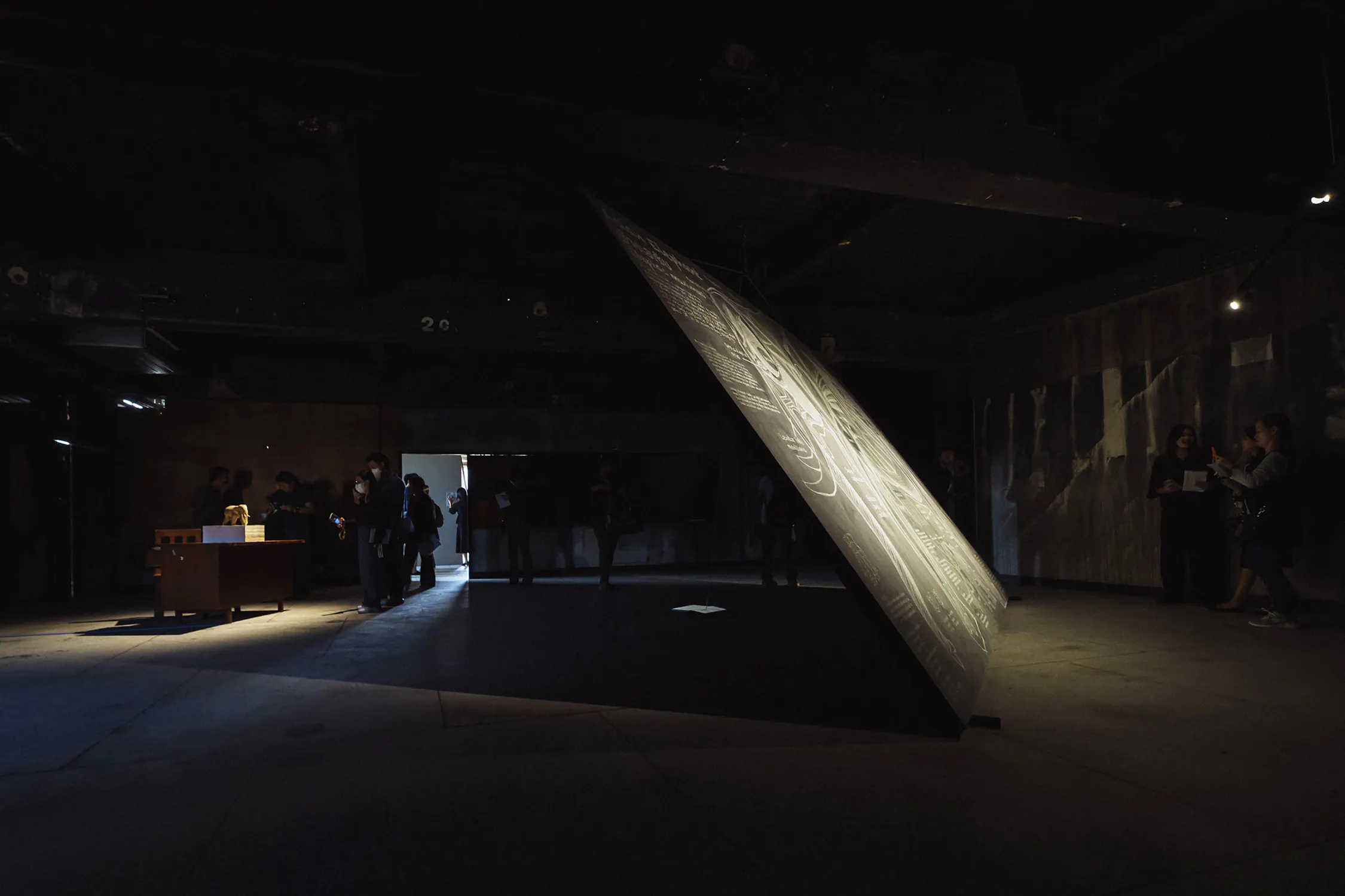 A large, tilted, rectangular monolith displays graphic illustrations and text in a dark room. Nearby, visitors stand near a small display table and observe the installation.