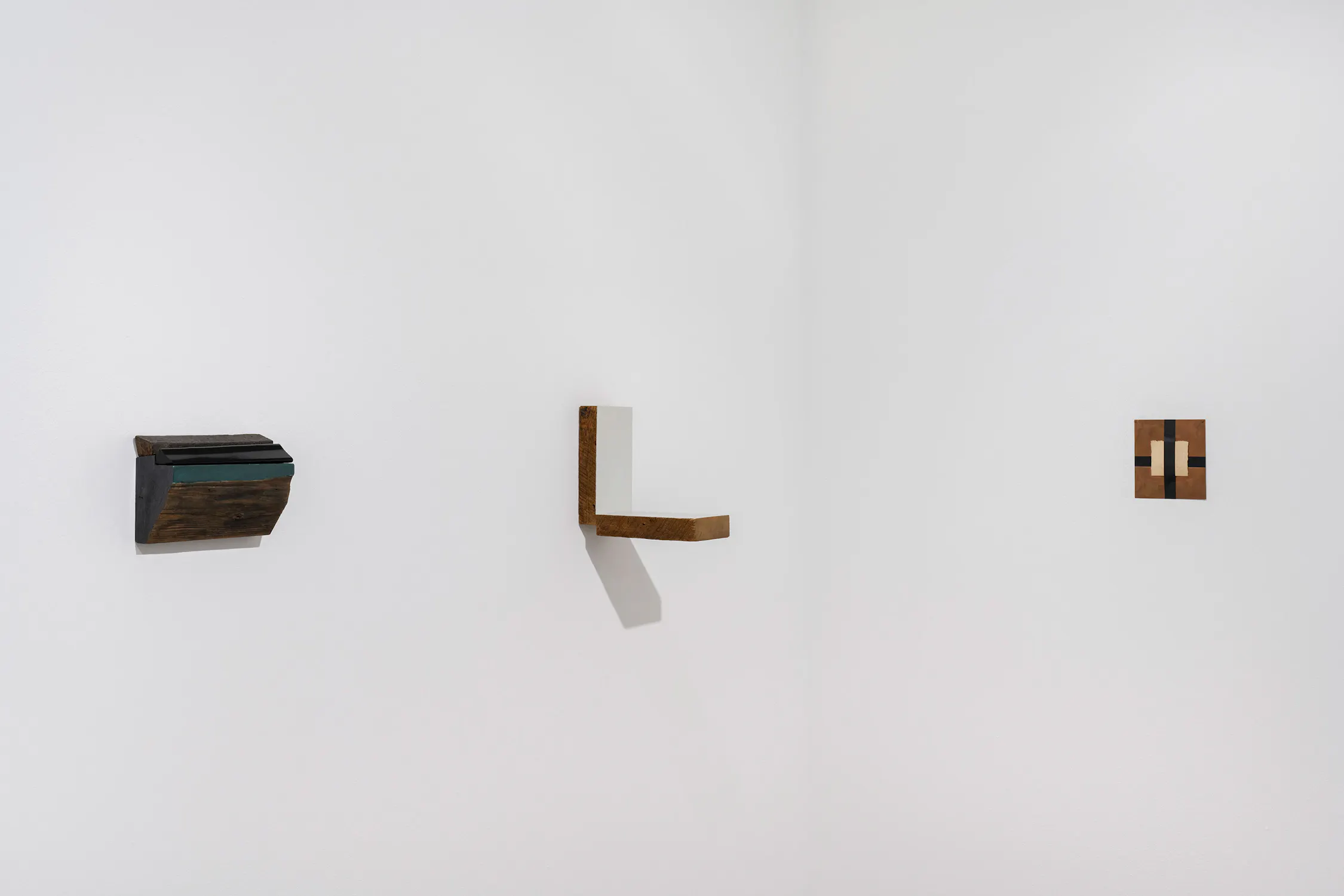 Three abstract, geometric wooden sculptures by Bill Walton are mounted on a plain white gallery wall. From left to right, they include a dark, multi-layered block, an L-shaped shelf, and a small, rectangular panel with light and dark inlaid wood patterns.