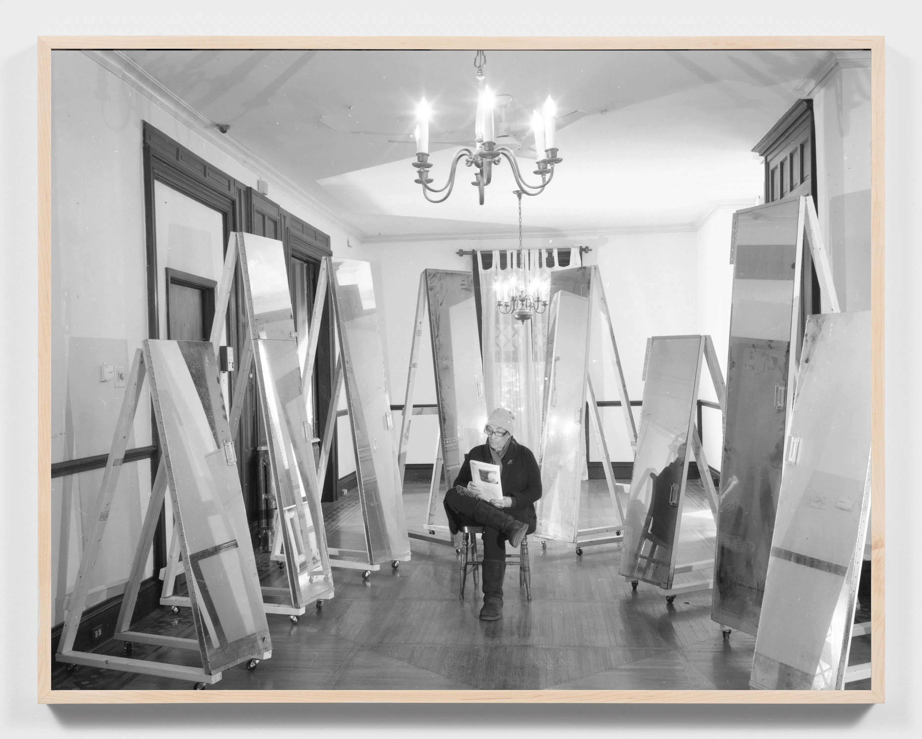 A person sits on a chair in the centre of a room, reading a book, surrounded by tall, rectangular mirrors on wheeled stands. The room has wood panelling, large windows, and a chandelier hanging from the ceiling. The photo is black and white.