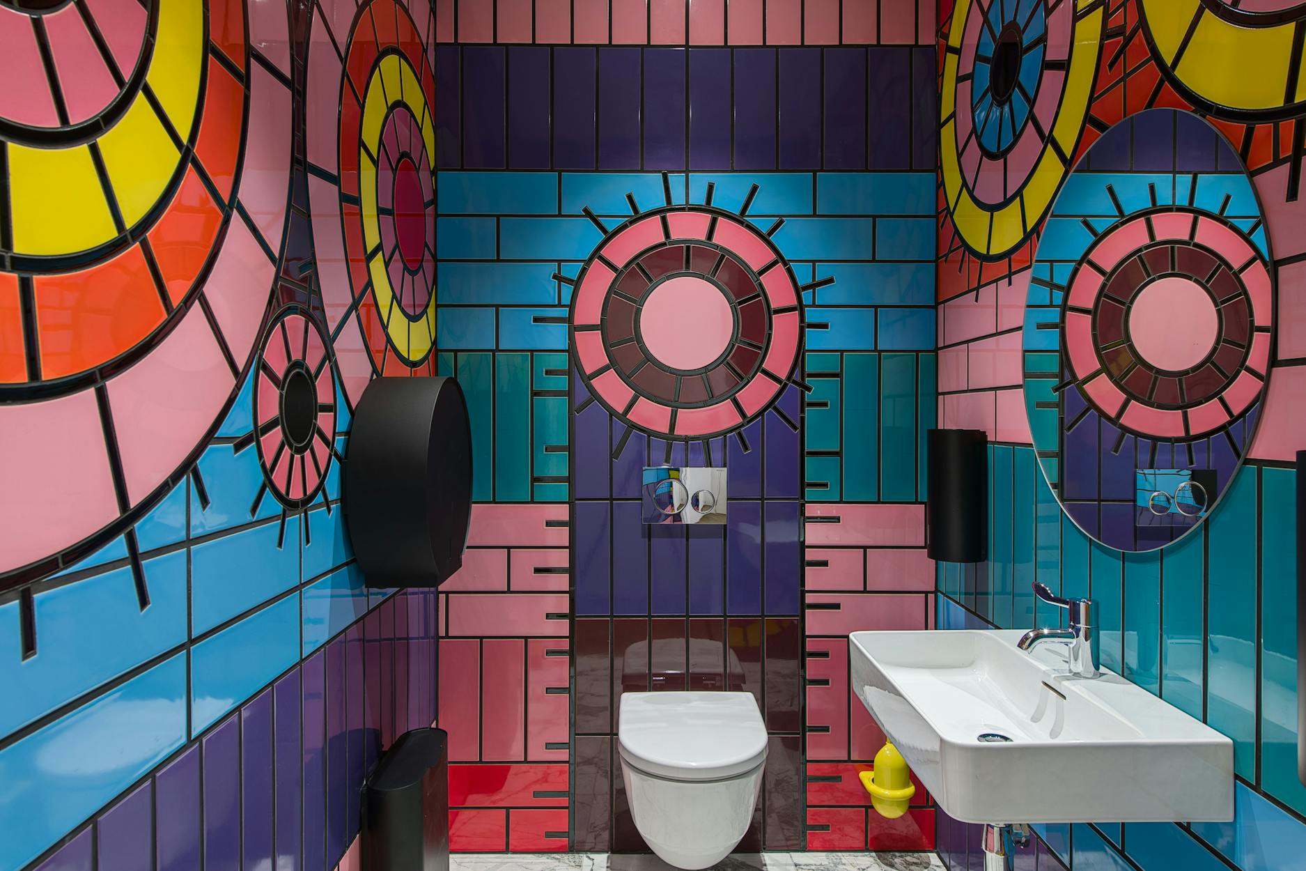 A bathroom with colourful, geometric tiled walls featuring concentric circle patterns in pink, yellow, red and blue. There is a wall-mounted toilet, white sink, mirrors and black bathroom fixtures.