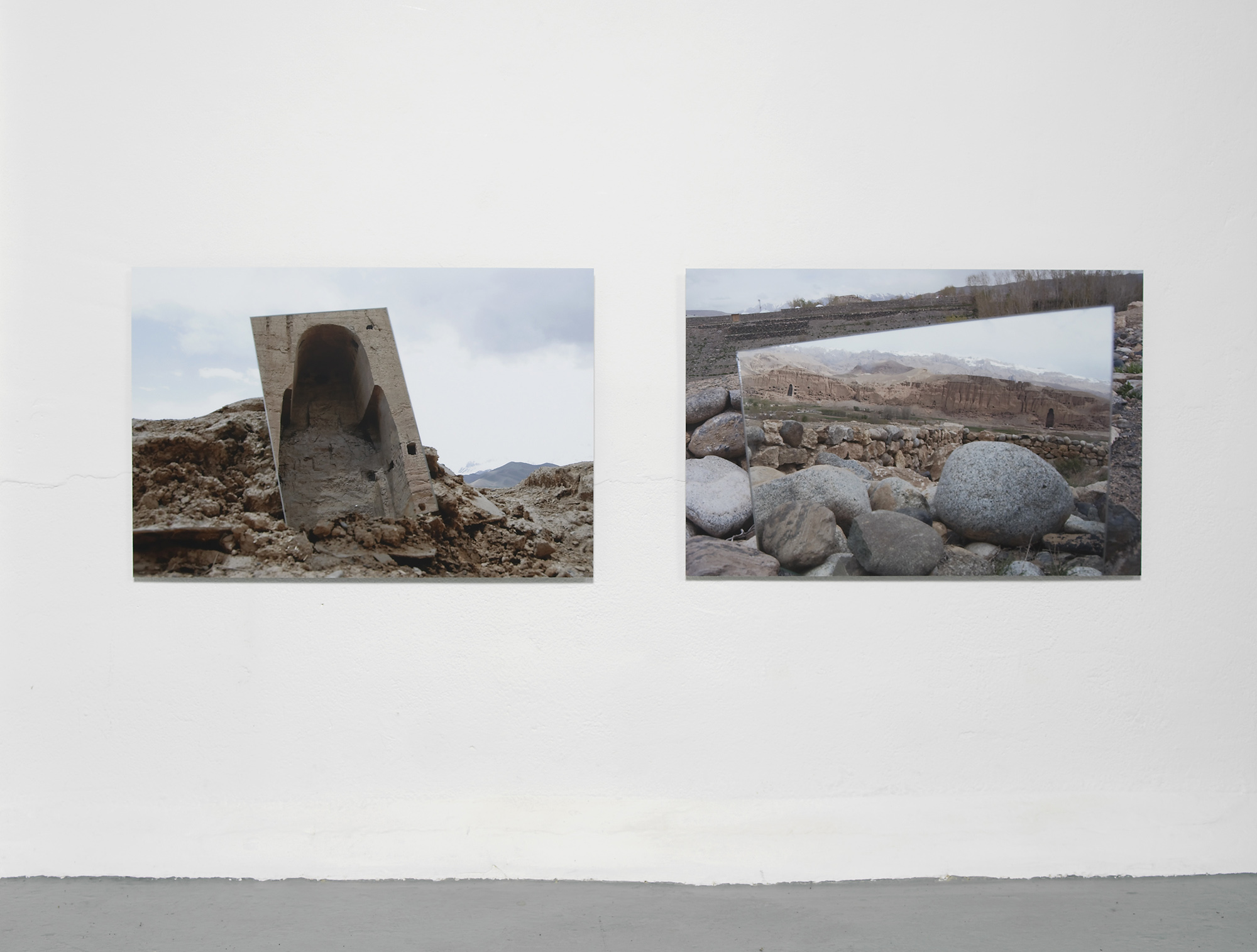 Two photographs hang on a white wall. The left shows a large, weathered, concrete structure among rocks. The right photo displays a landscape reflected in a rectangular mirror resting on stones.