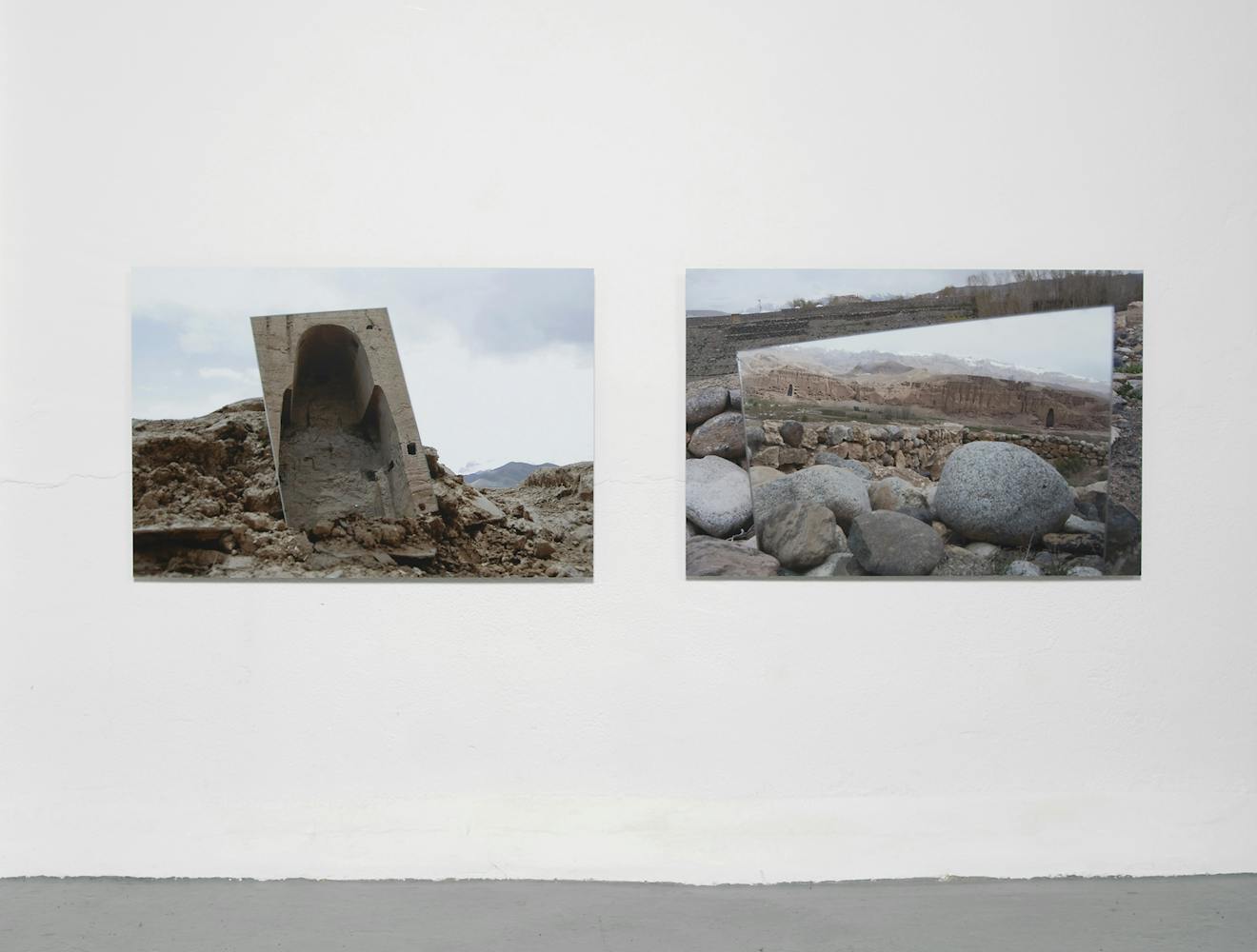 Two photographs hang on a white wall. The left shows a large, weathered, concrete structure among rocks. The right photo displays a landscape reflected in a rectangular mirror resting on stones.