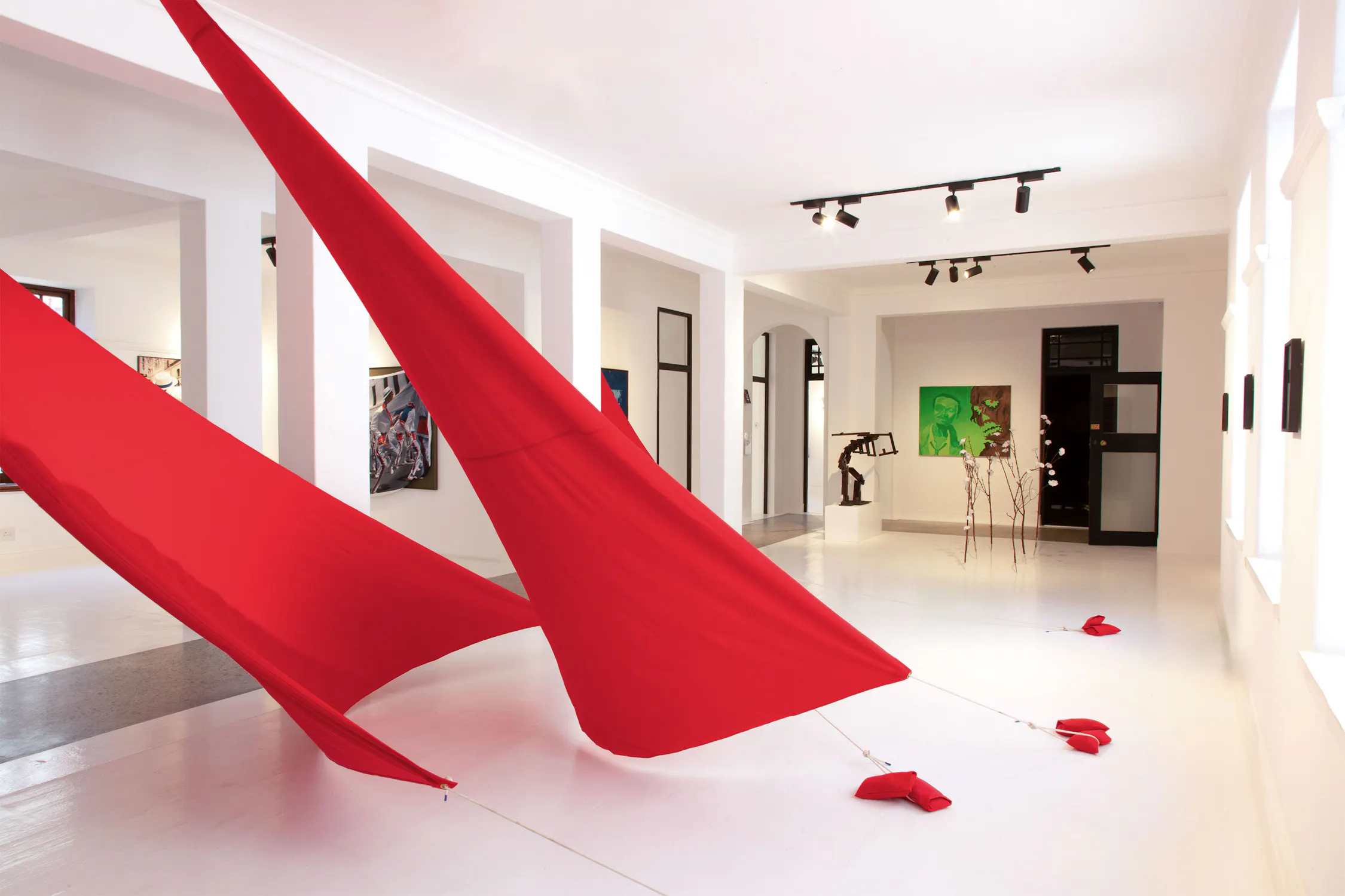 A contemporary art gallery displays red fabric installations stretched diagonally across a white floor, anchored at four points. Paintings and sculptures are visible in the background. The room is bright with white walls, large windows, and spotlights on the ceiling.