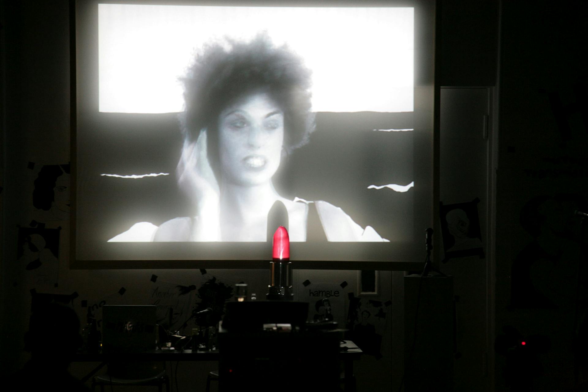 a black and white film is projected onto a screen in a darkened room. A performer is shown on screen with curly hair. A giant novelty red lipstick prop obscures part of the screen.