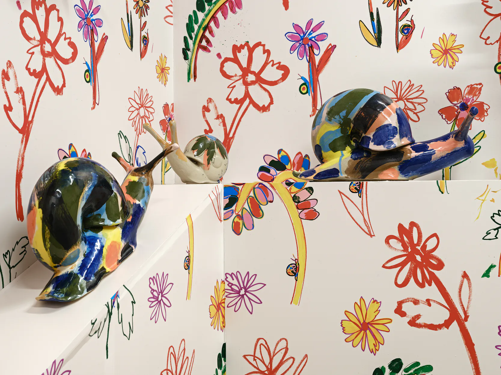 Two large, colourful ceramic snails by Supergroup are displayed against white walls covered with hand-drawn, multicoloured flowers, leaves, and abstract shapes, creating a vibrant, playful scene.