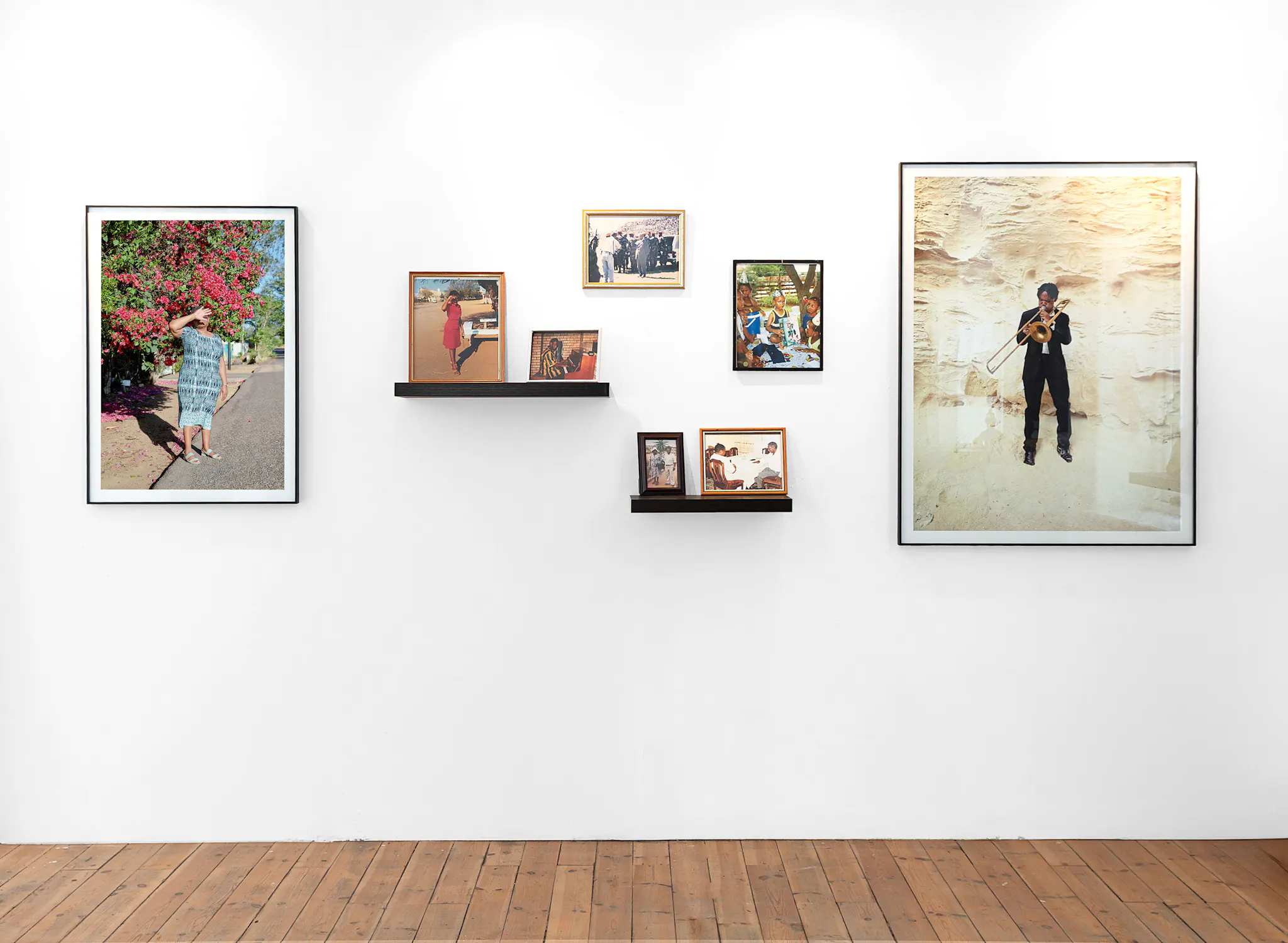 A white gallery wall displays eight framed photos, some on small black shelves. Images show people posing, dancing, and playing trumpet. The floor is wooden, and the overall scene is bright and well-lit.