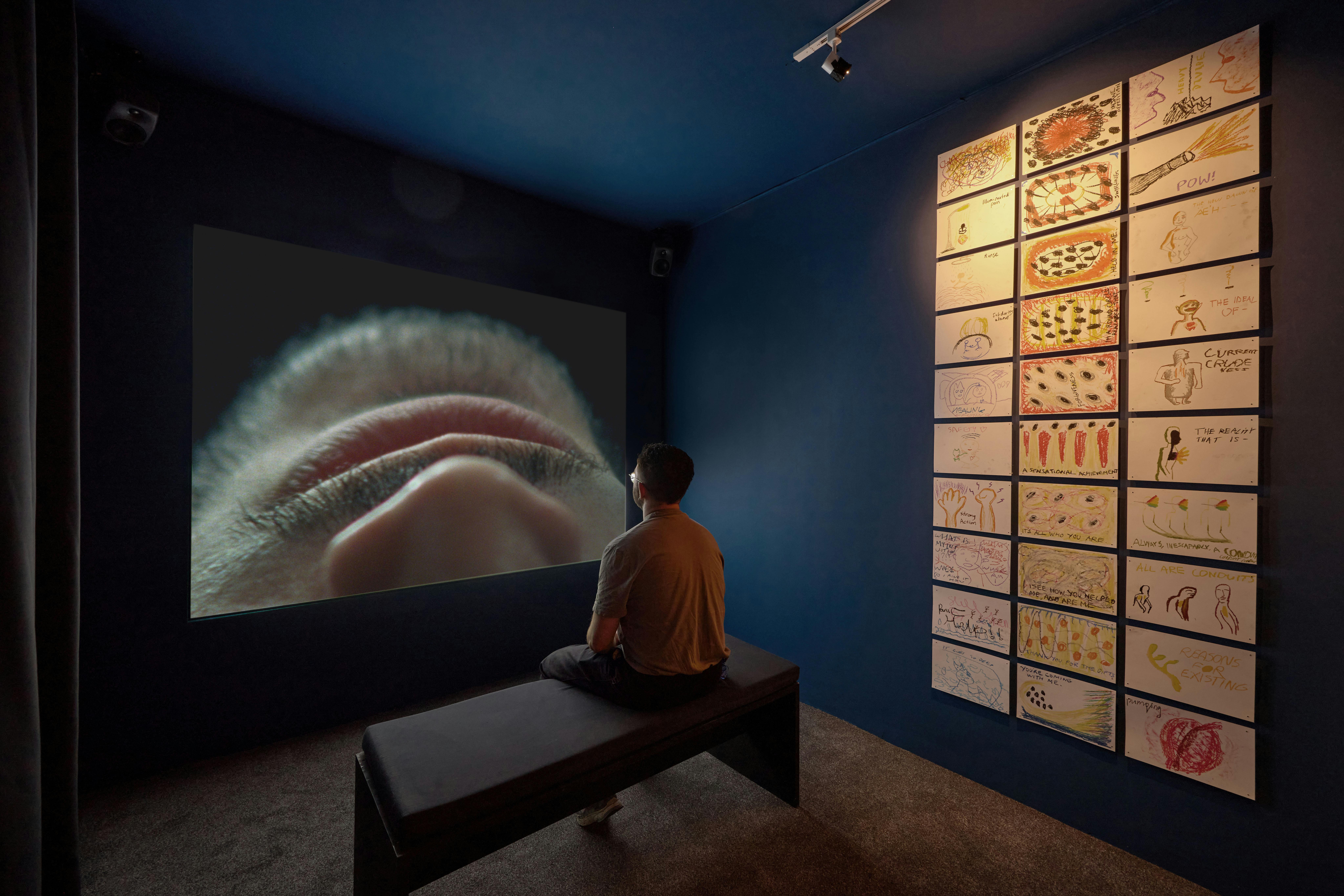 A person sits in a dark room on a bench, facing a large screen displaying a close-up of a nose and lips. To the right, a wall displays a grid of drawings with various colors and designs.