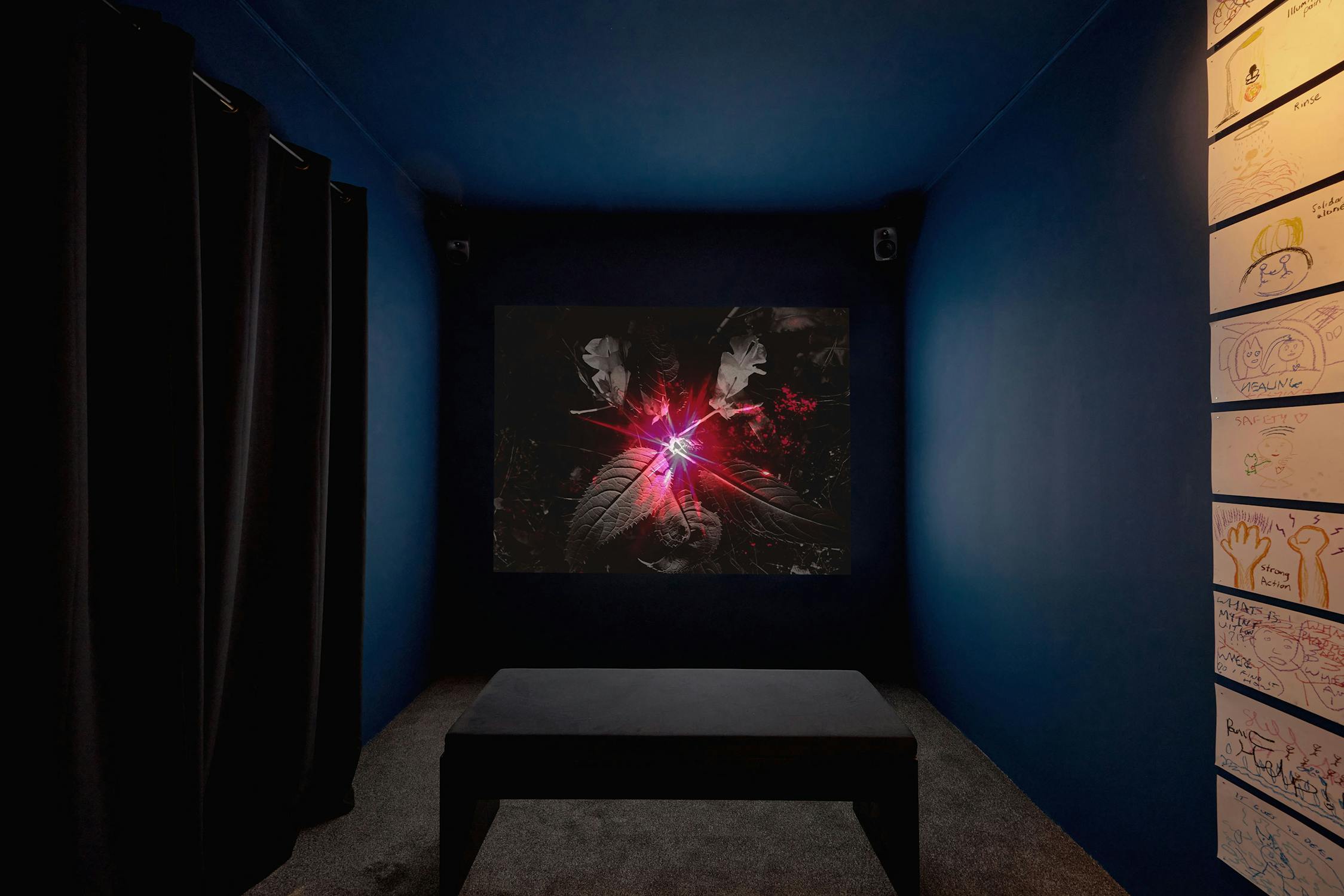 A dark room with blue walls displays a screen showing a surreal image with red light and abstract forms. A black bench is in the centre. The wall on the right features a grid display of drawings.