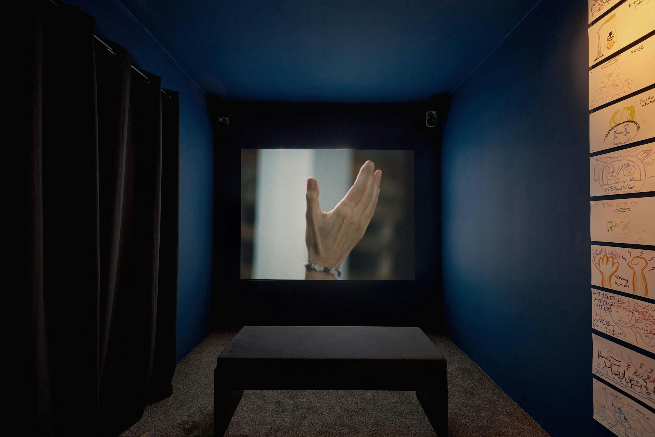 A dark room with a projector screen displaying an image of an outstretched hand. The walls are blue, and there's a bench in the centre. Drawings are displayed on the right wall. Thick curtains cover part of the left wall.