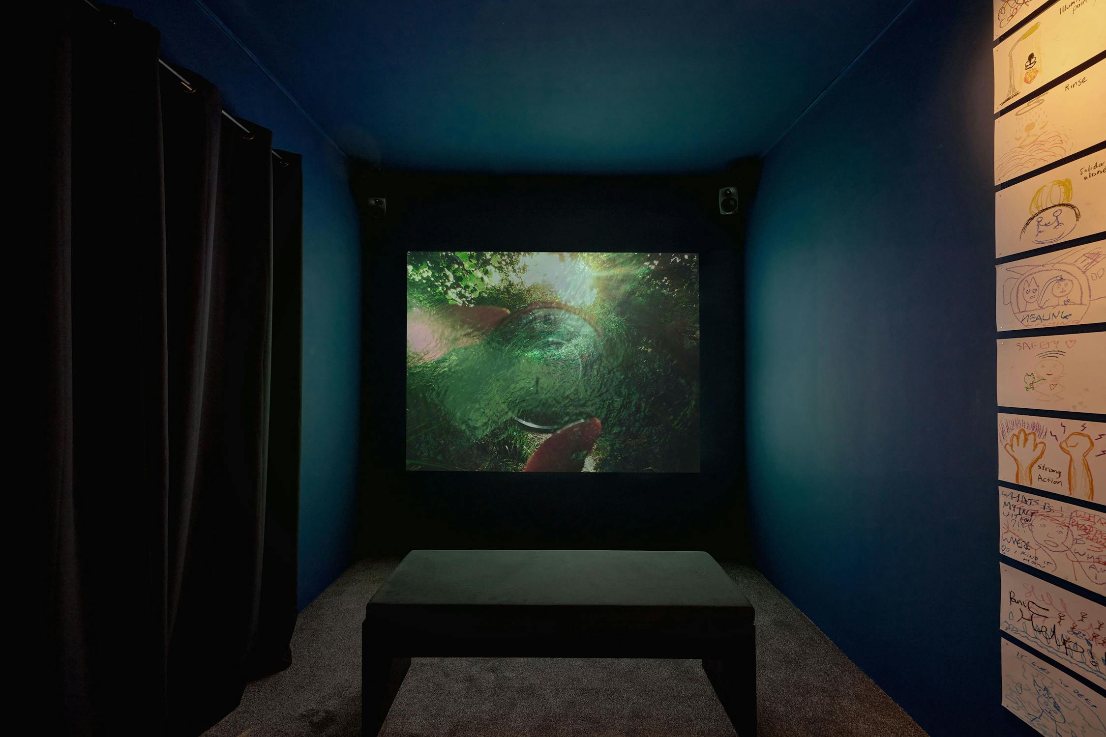 A dark room features a large screen displaying a green, nature-themed video. A black bench is in the centre. The wall on the right features a grid display of drawings.