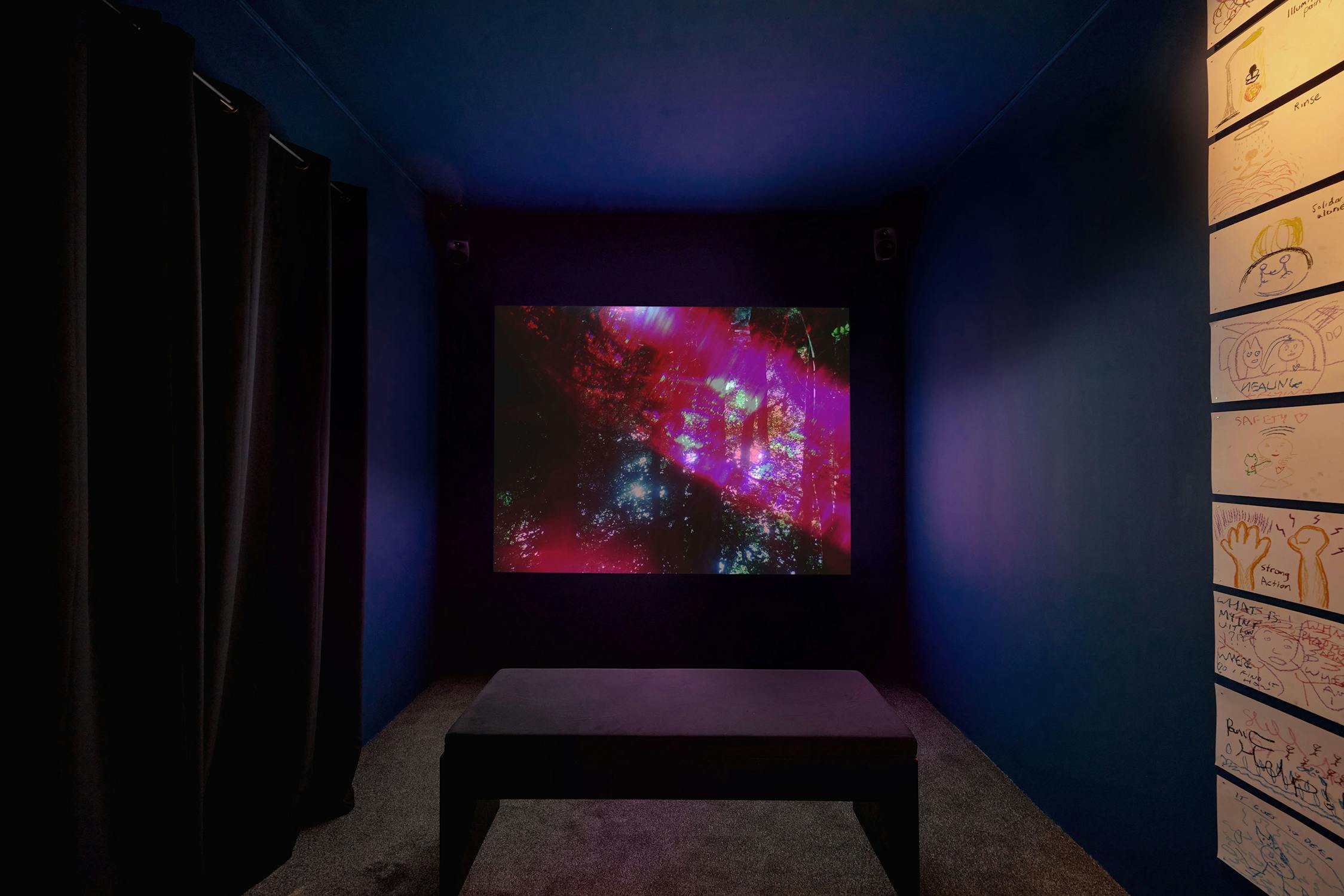 A dark room with a bench facing a large screen displaying abstract, colorful patterns. Black curtains are on the left, and the right wall features a grid display of drawings and sketches. 