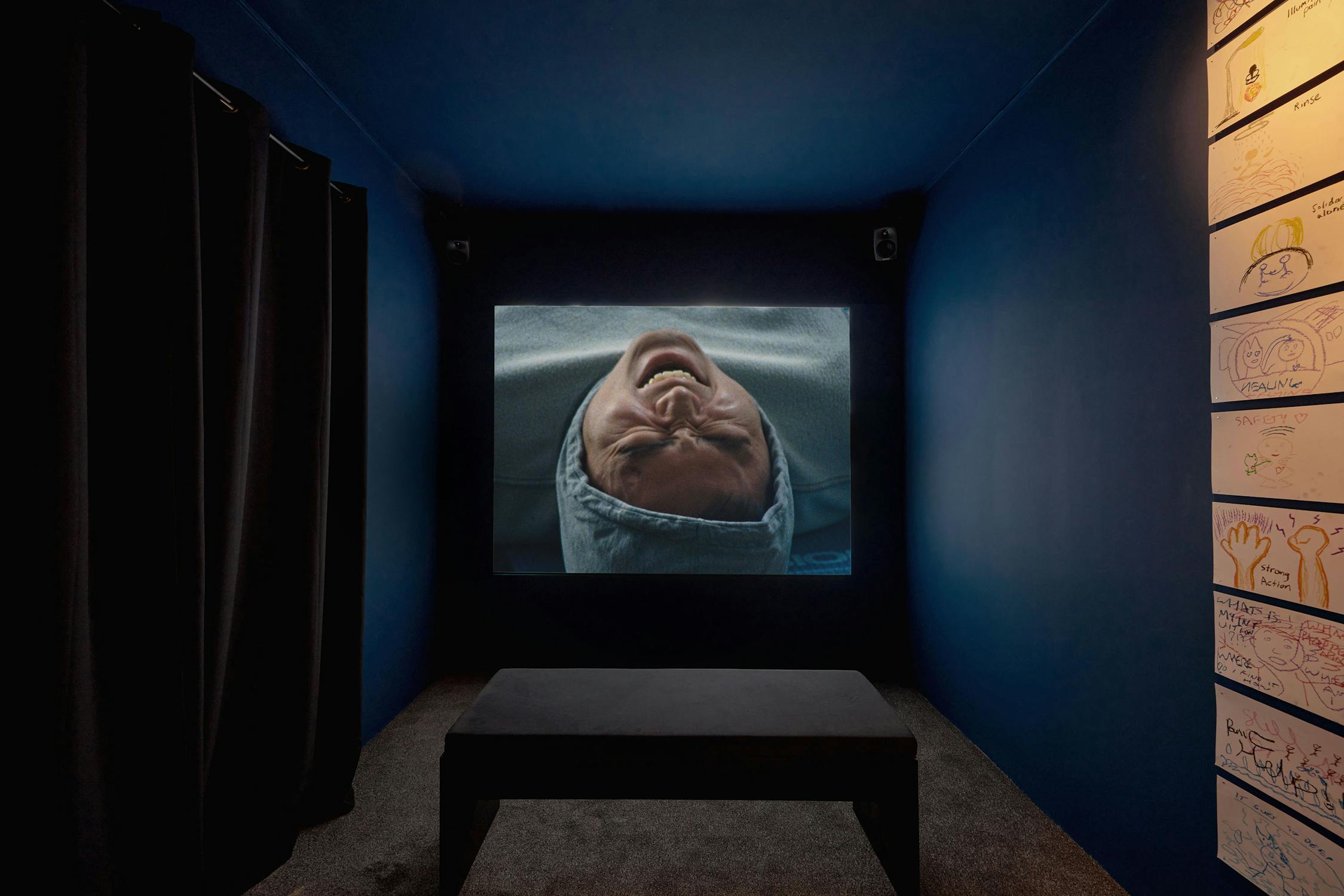 A small dark room with a large screen displaying an image of a person in a hood. The person appears to be grimacing. There is a bench in the centre. The walls have drawings displayed on the right, and to the left, there is a curtain covering the doorway.