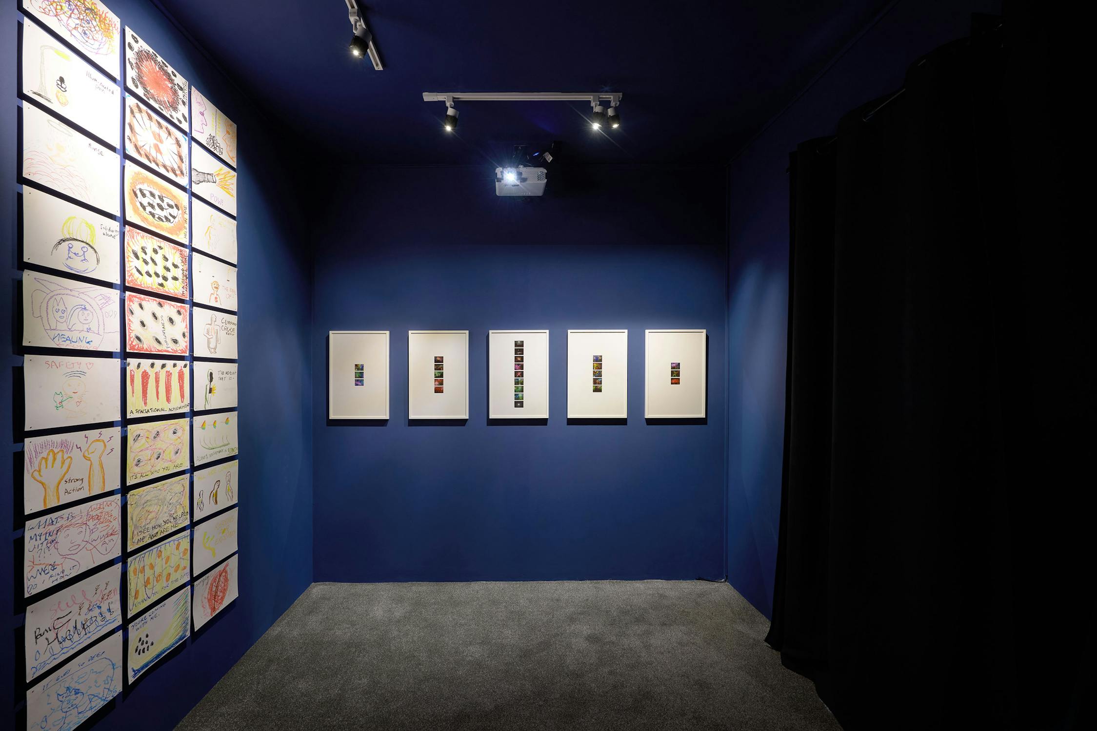 A small art gallery room with dark blue walls features framed abstract artworks. One wall displays a grid of colourful drawings, while the opposite wall has five framed pieces. Overhead lighting illuminates the exhibits. Black curtains are visible to the right.