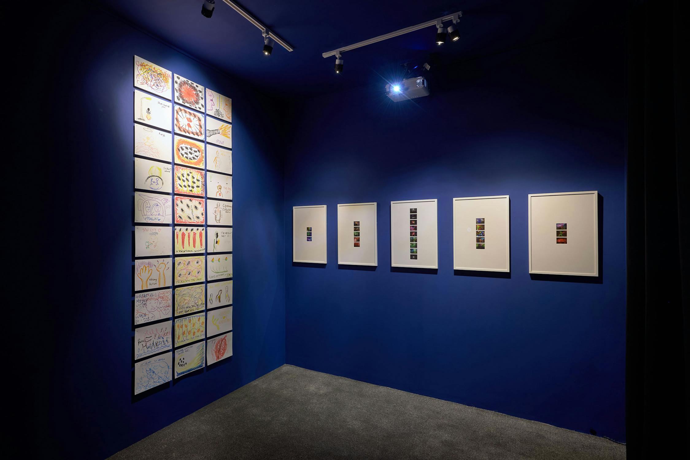A deep blue gallery wall displays a grid of small, colourful paintings on the left and five framed art pieces featuring vertical rows of vivid squares on the right. The space is dimly lit with spotlights.