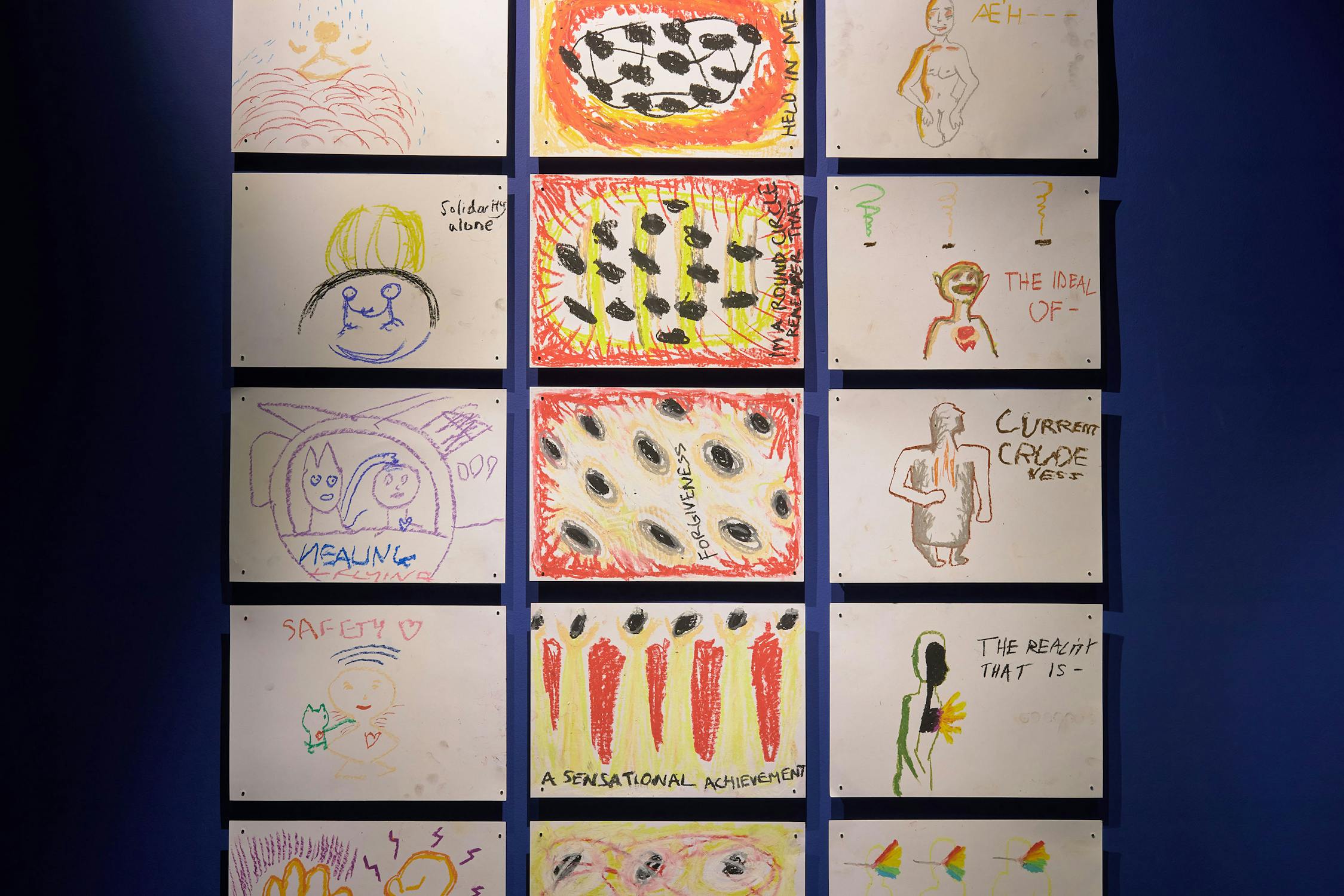 A grid of various colourful drawings on a blue wall. The drawings include abstract shapes, faces, text, and objects like a pizza and a figure holding a balloon. Some words, such as "Solidarity" and "Safety," are visible on the drawings.