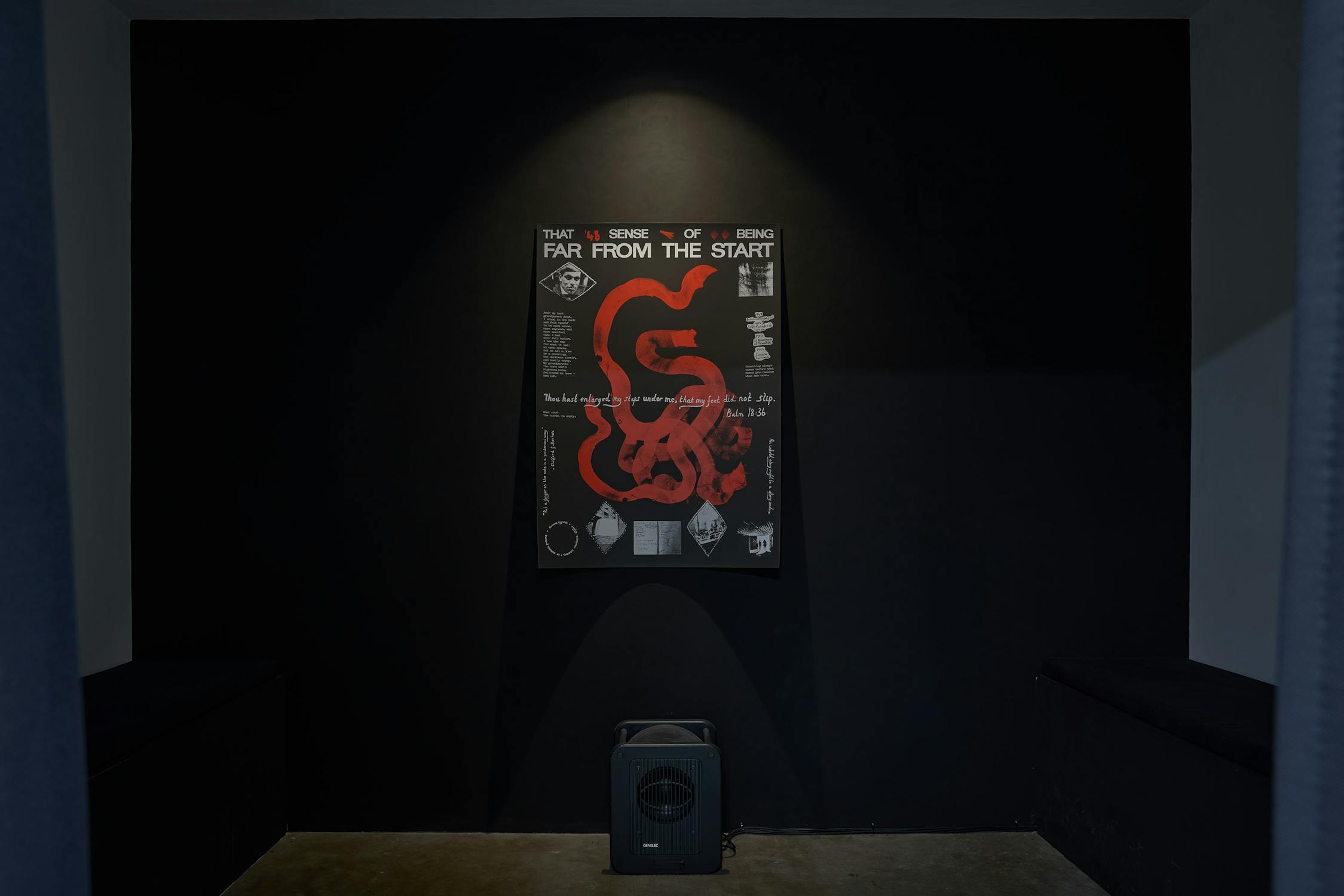 A dark room with a spotlight illuminating a poster by Jay Bernard on the wall. The poster features abstract red and white designs with text. Below the poster is a black cube-shaped object. The walls and surroundings are painted black.