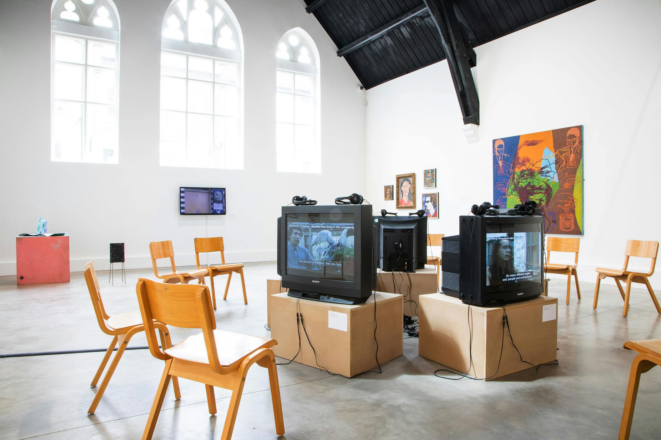 An art gallery with rows of wooden chairs facing several CRT televisions on cardboard boxes. The TVs display various images. The room has high windows with arched tops and artworks on white walls.