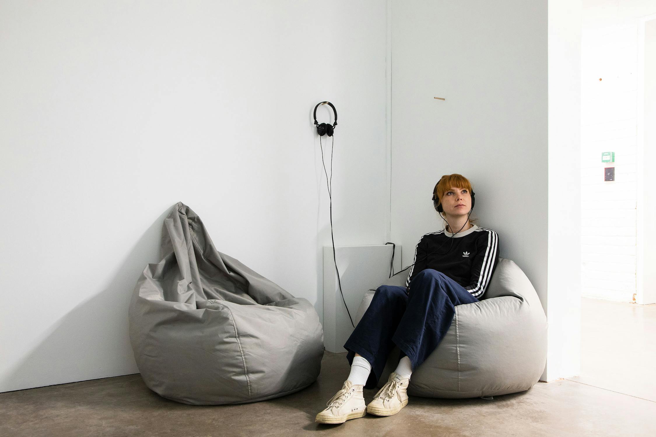A person with headphones sits on a gray beanbag in a minimalist room with white walls. Another beanbag is nearby, and headphones hang on the wall. The person wears casual clothes and appears relaxed.