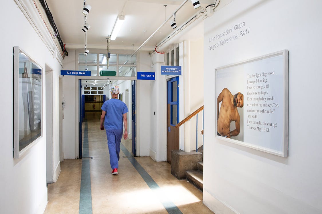 A person in scrubs walks down a hospital hallway. The walls display art, including a framed photograph of a shirtless man bending  and stretching. Overhead signs indicate directions to various departments like IT Help Desk and MRI/CT Scans.