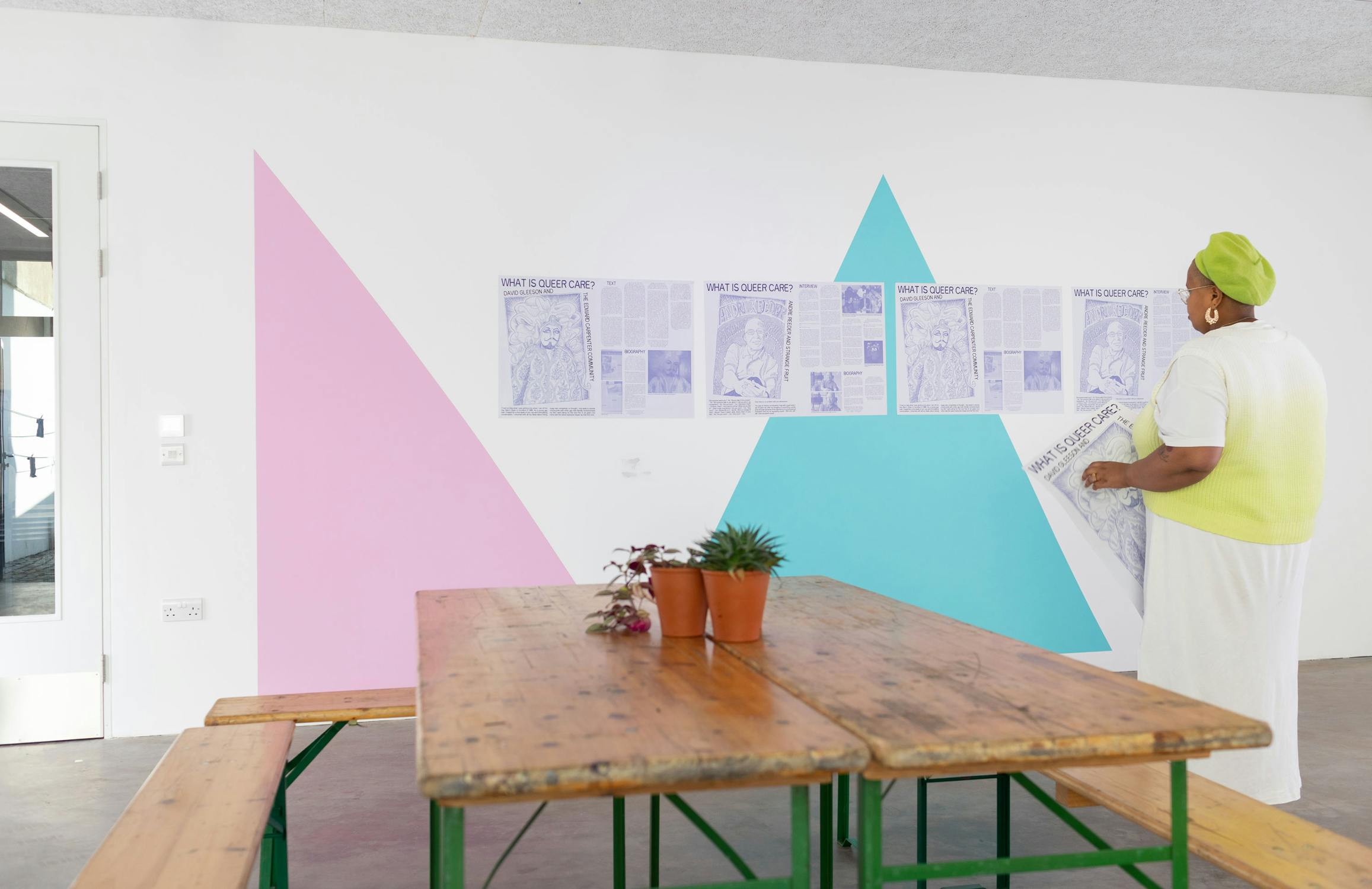 A person in a green beret and gradient dress looks at newspaper articles displayed on a wall with pink and blue geometric shapes. A wooden table with potted plants is in the foreground.