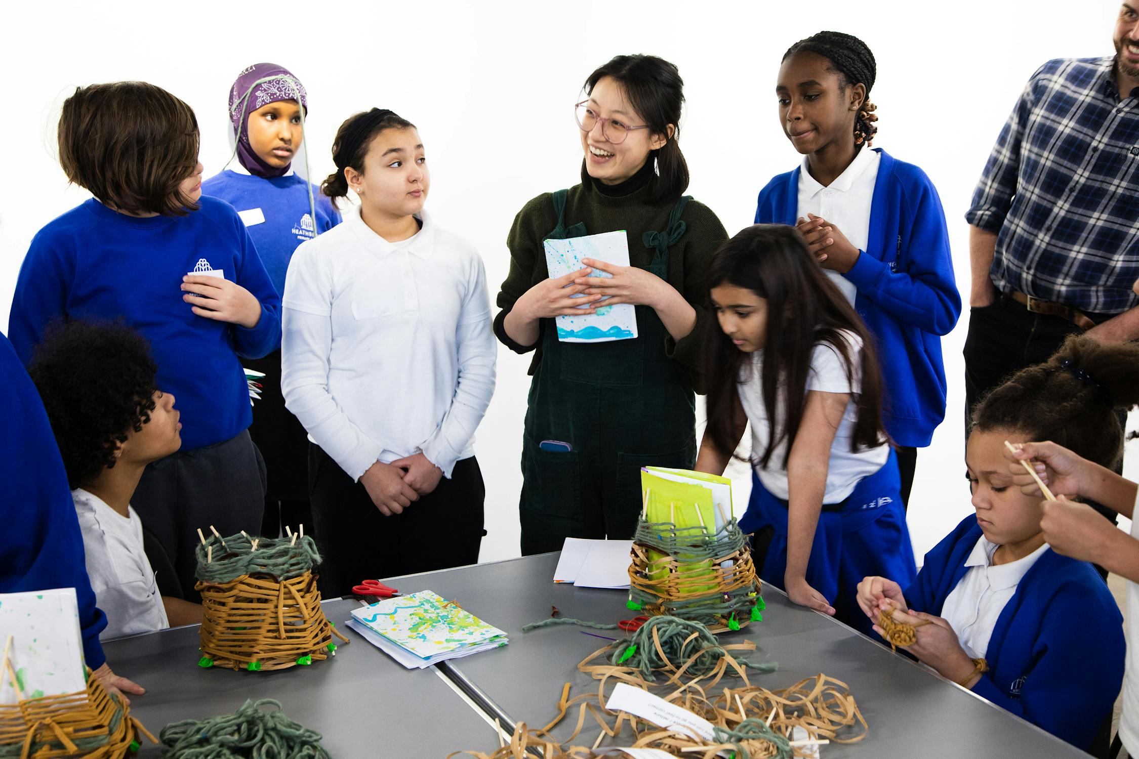 A group of children from Heathbrook Primary School in blue uniforms gather around a table, engaged in an art activity with yarn and craft supplies. Art coordinator Amy Leung holds a book, leading the activity. Some children are focused on their crafts, while others look at the adult attentively.