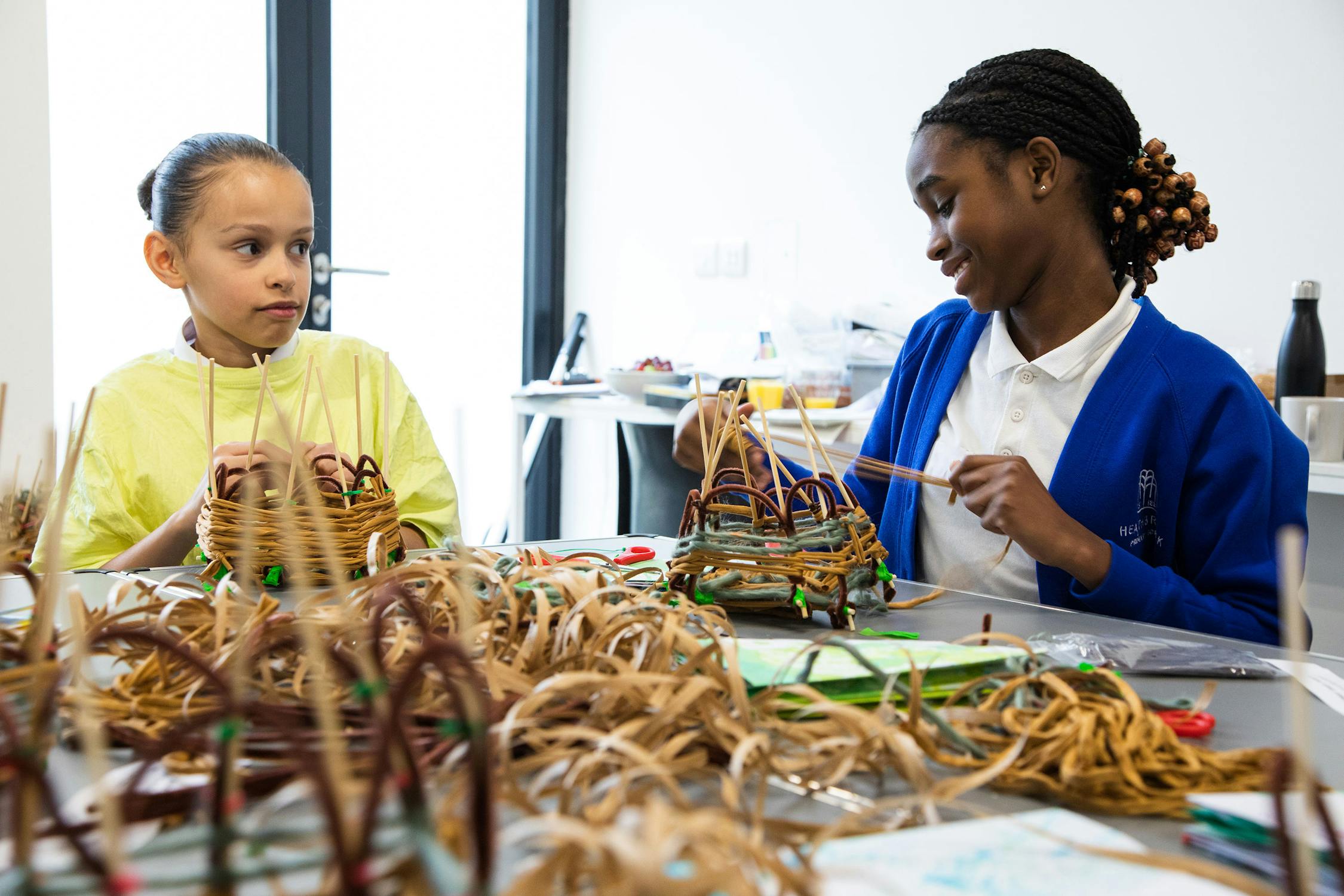 Two children sitting at a table engaged in a craft activity with natural materials. One child in a yellow shirt is looking at the other, who is smiling and wearing a blue sweater. The table is covered with woven pieces and tools.