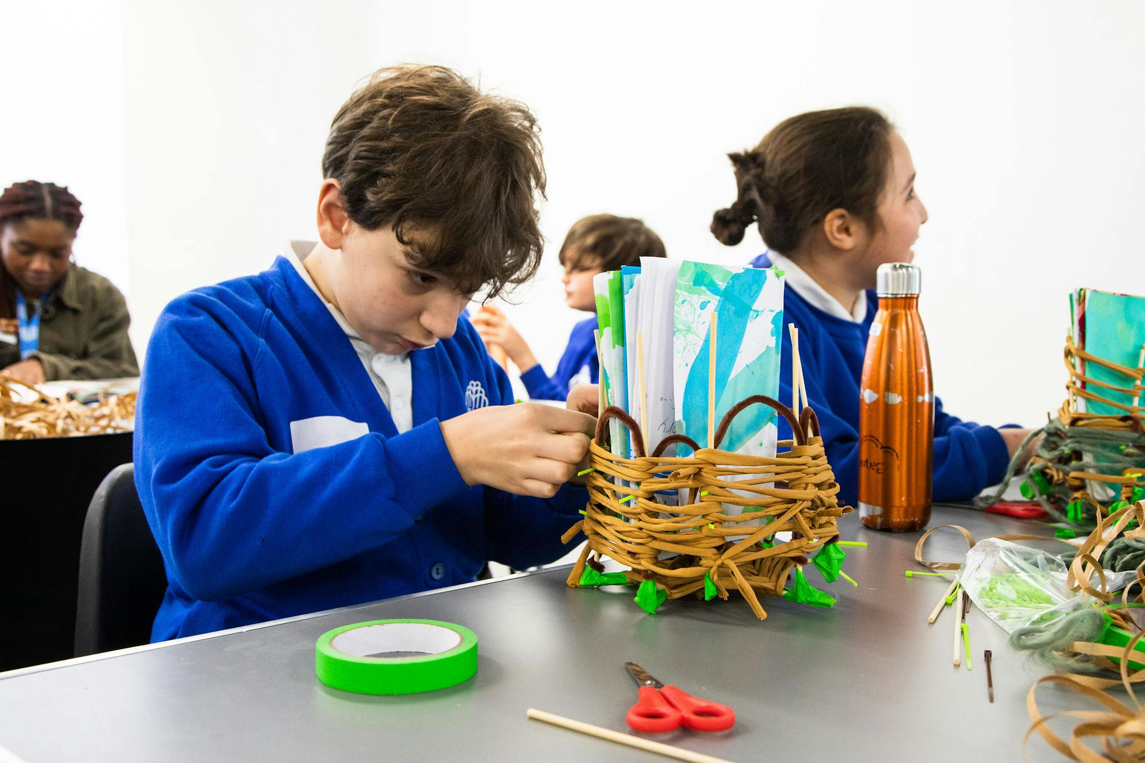 A group of children wearing blue uniforms are seated at a table, engaged in crafting with woven materials. One boy focuses on a basket, while others work and watch. Supplies like scissors and tape are visible on the table.