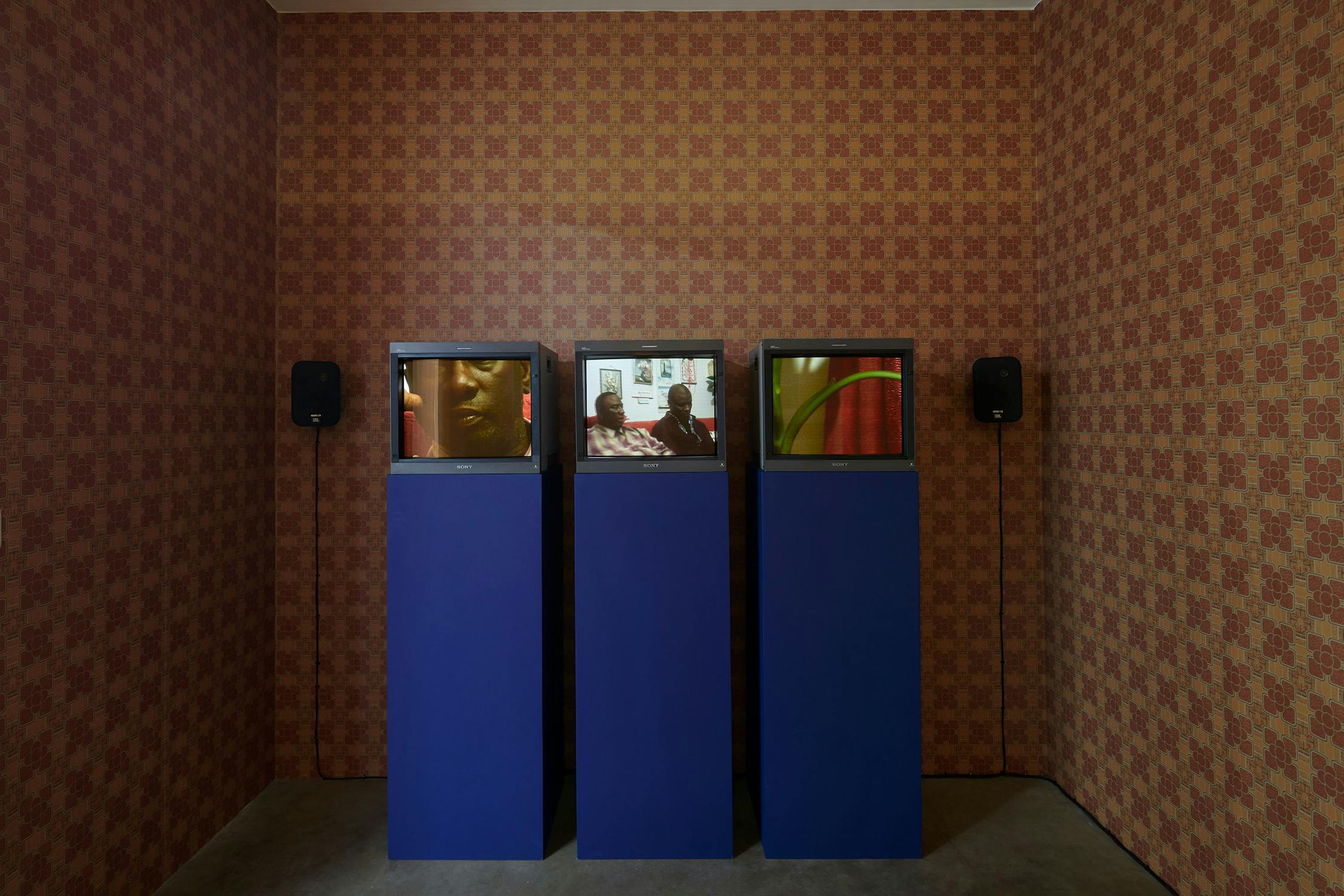 Three vintage televisions on blue pedestals display different scenes against a brown, patterned wallpaper background. The televsions show a three-channel film by Holly Graham. Each screen shows distinct images, one featuring a close-up, another a portrait, and the third with abstract shapes.