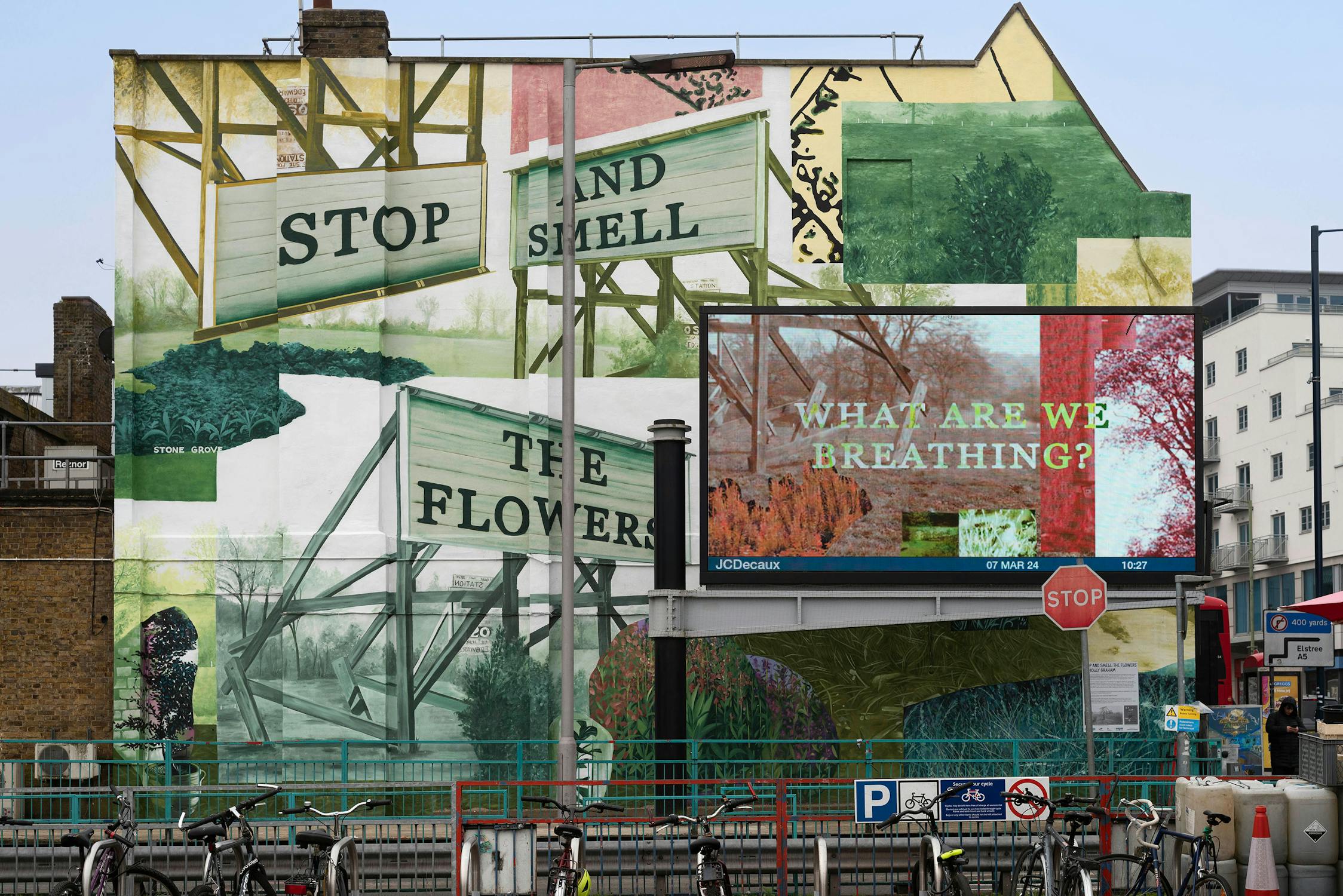A mural by Holly Graham on a brick building shows signs saying "Stop and smell the flowers" among green foliage. Next to it, a billboard asks, "What are we breathing?" Bicycles are parked in front, along with a stop sign and parking symbols.