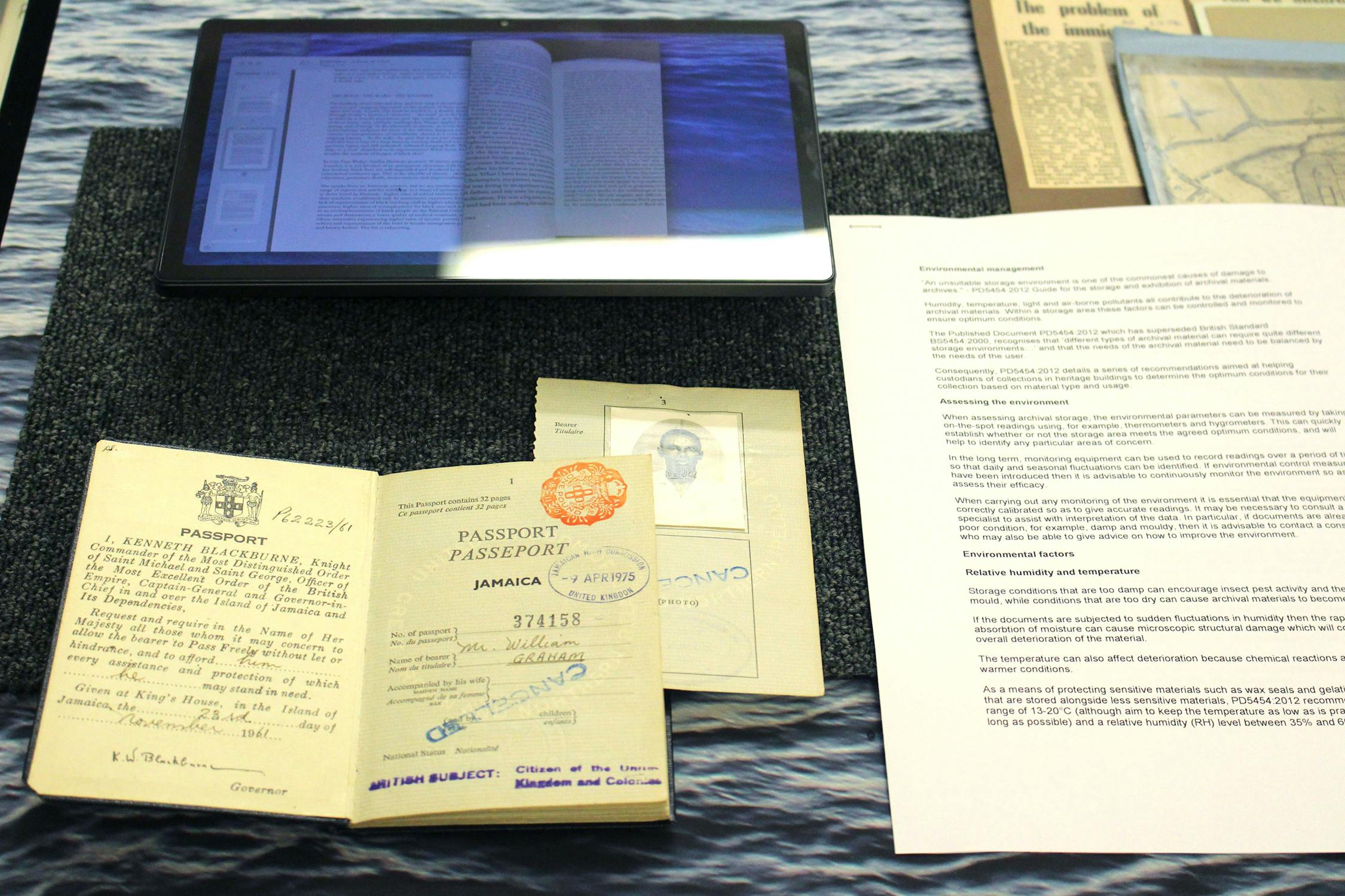 An open passport with a stamp and handwritten notes is displayed. Next to it is a typewritten letter on white paper. A tablet showing a digital document is in the background, all placed on a blue textured surface.