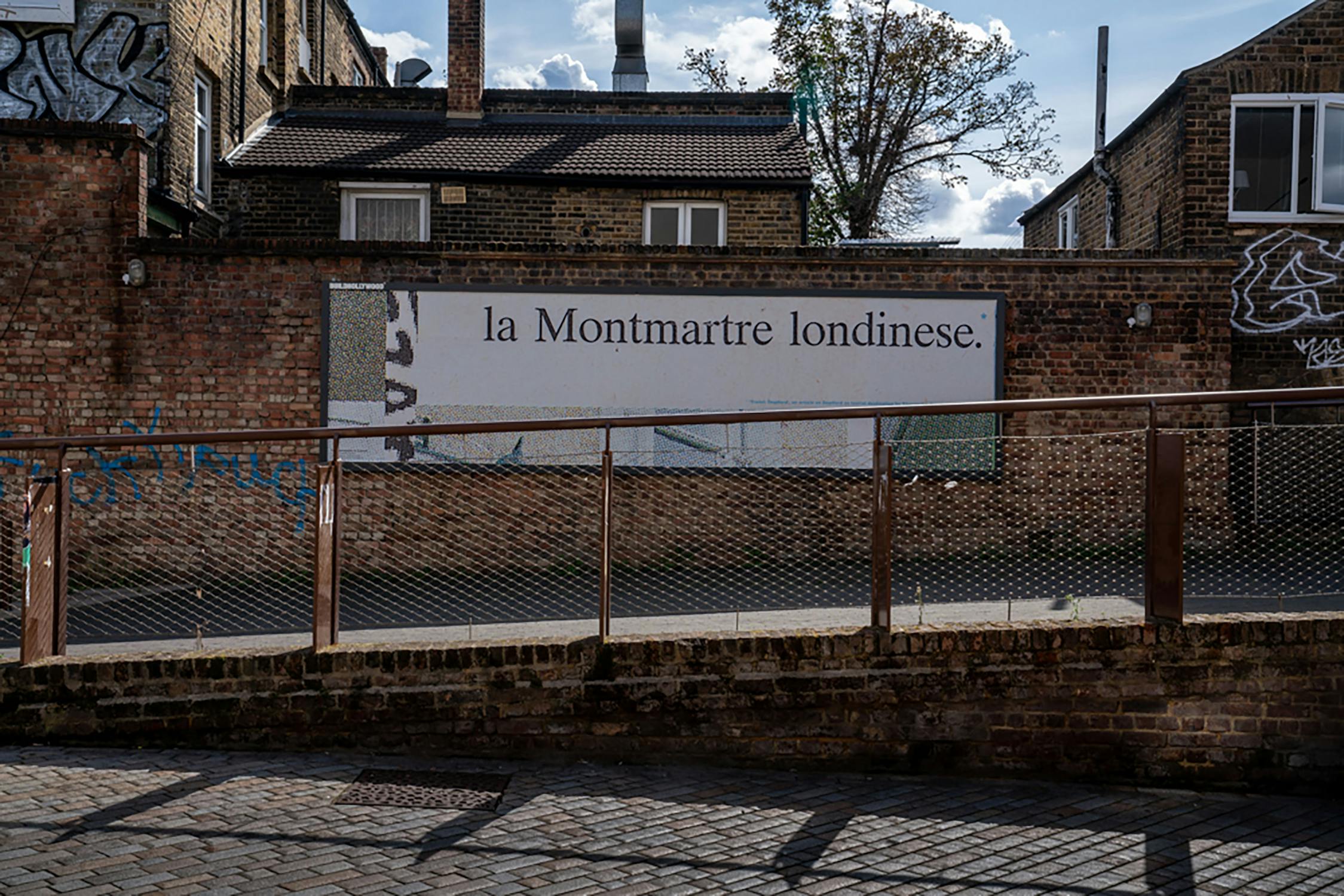 A cobblestone street leads to a brick wall with a large sign reading "la Montmartre londinese." The area has a mix of industrial and residential buildings, some with graffiti. A partly cloudy sky is visible above.