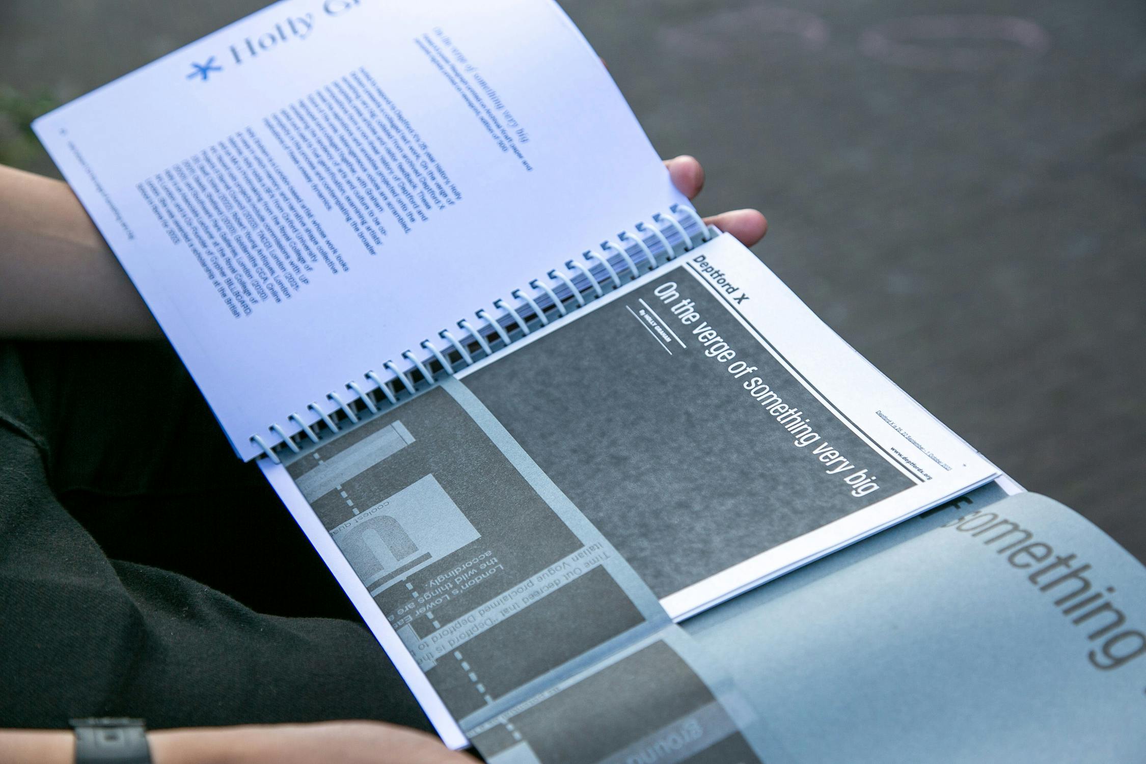 Person holding an open spiral-bound book with pages titled "On the verge of something very big" and visible text on other pages. The background shows a blurry outdoor surface.
