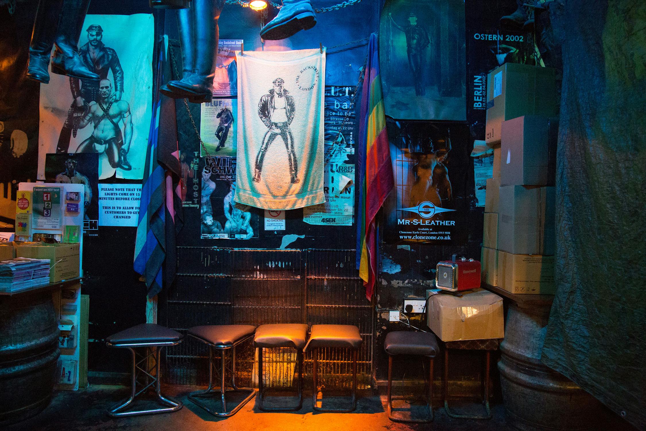 A dimly lit room with a collection of posters, including a prominent one of a person in leather attire. There are colourful flags, stacked boxes, and four stools arranged along the wall, creating a cosy, eclectic atmosphere.