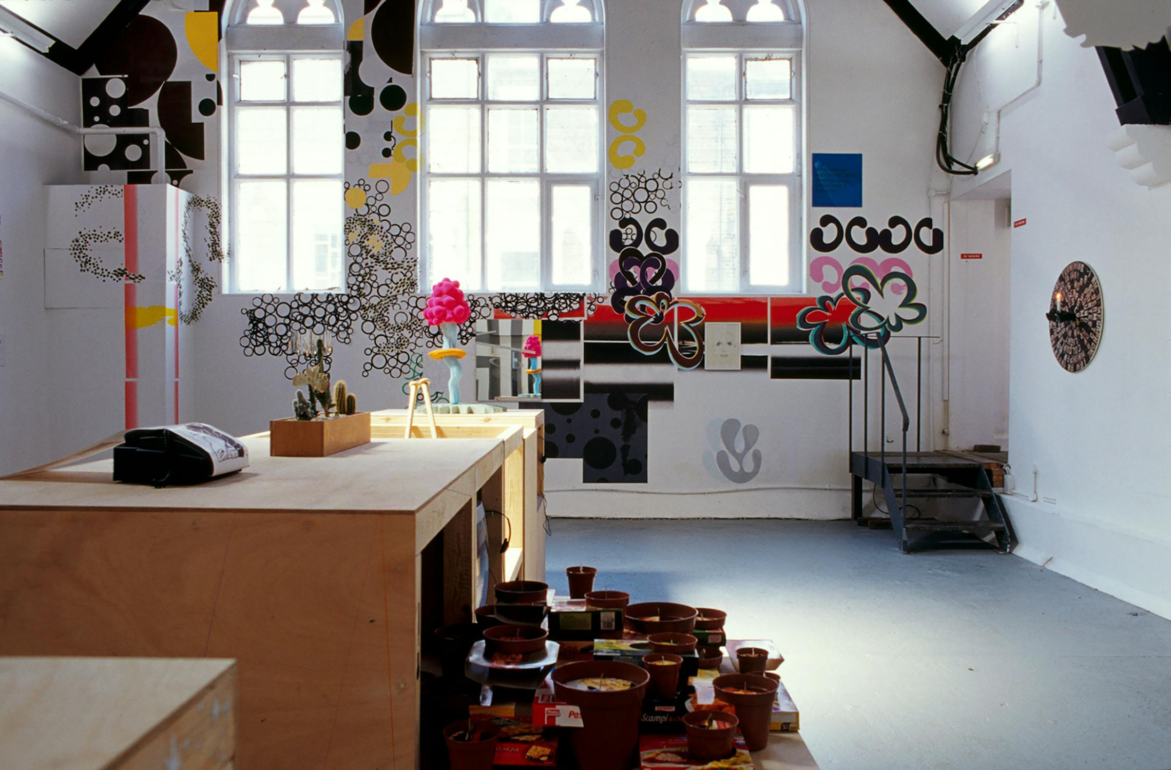 A brightly lit, modern room with decorative graffiti-style wall art, featuring abstract designs and colourful patterns. The room has a wooden counter with potted plants and assorted items. Large windows allow natural light to fill the space.