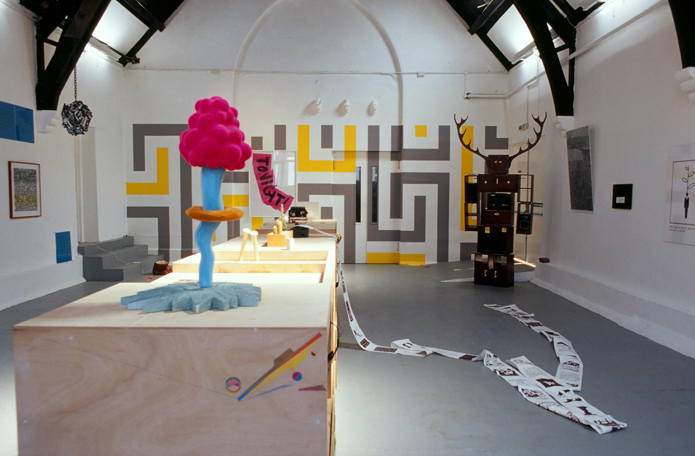 Installation art featuring wooden platforms with colourful abstract sculptures, including a pink cloud-like structure and blue geometric shapes. A maze-like grey and yellow mural is on the wall, and small shelves hold various objects. Papers trail across the floor.