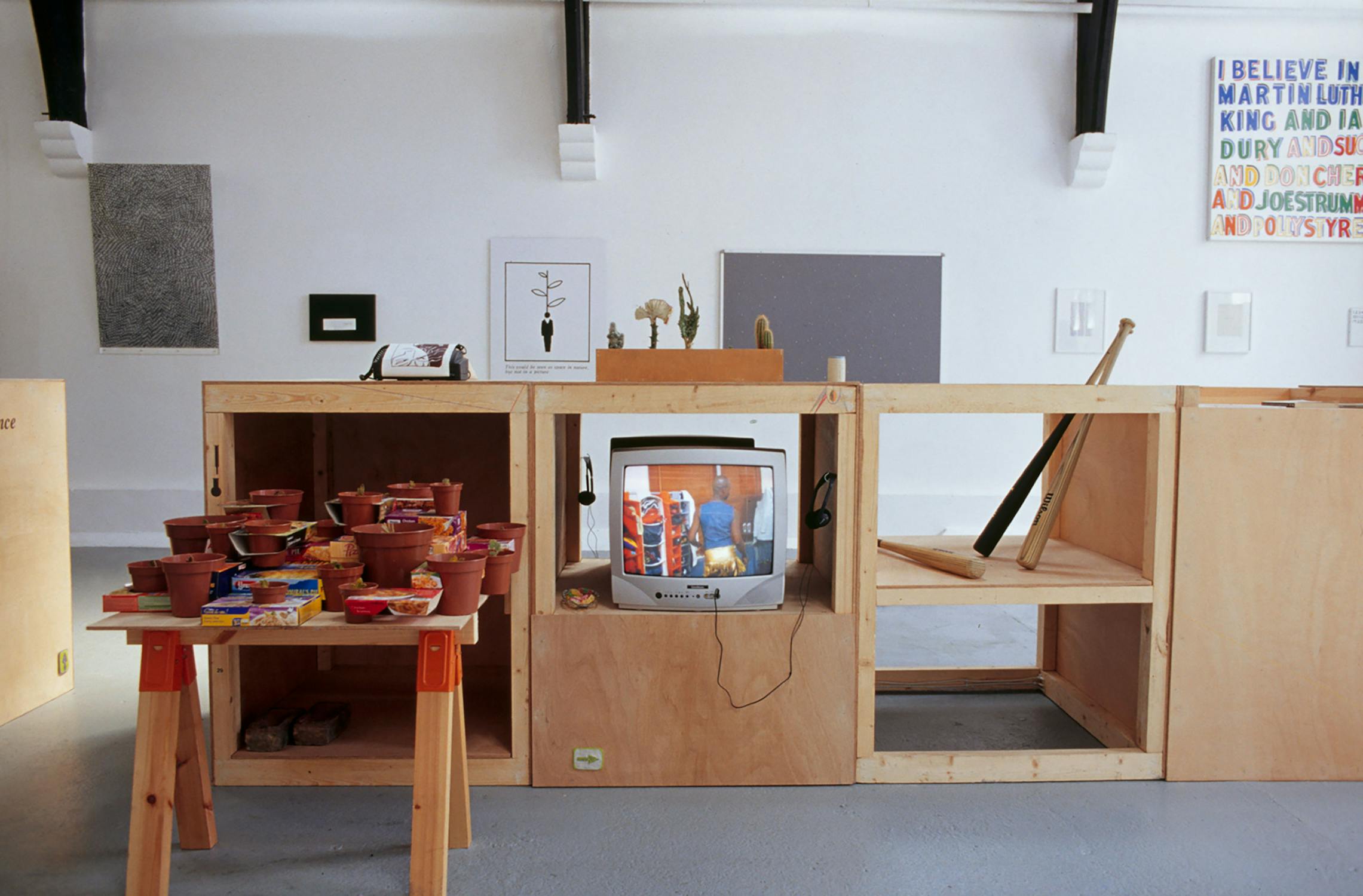 Art installation featuring a wooden structure with various objects: potted plants, a small TV displaying an image, and shelves. A table with colourful bowls is in the foreground. Abstract framed artworks hang on a white wall.