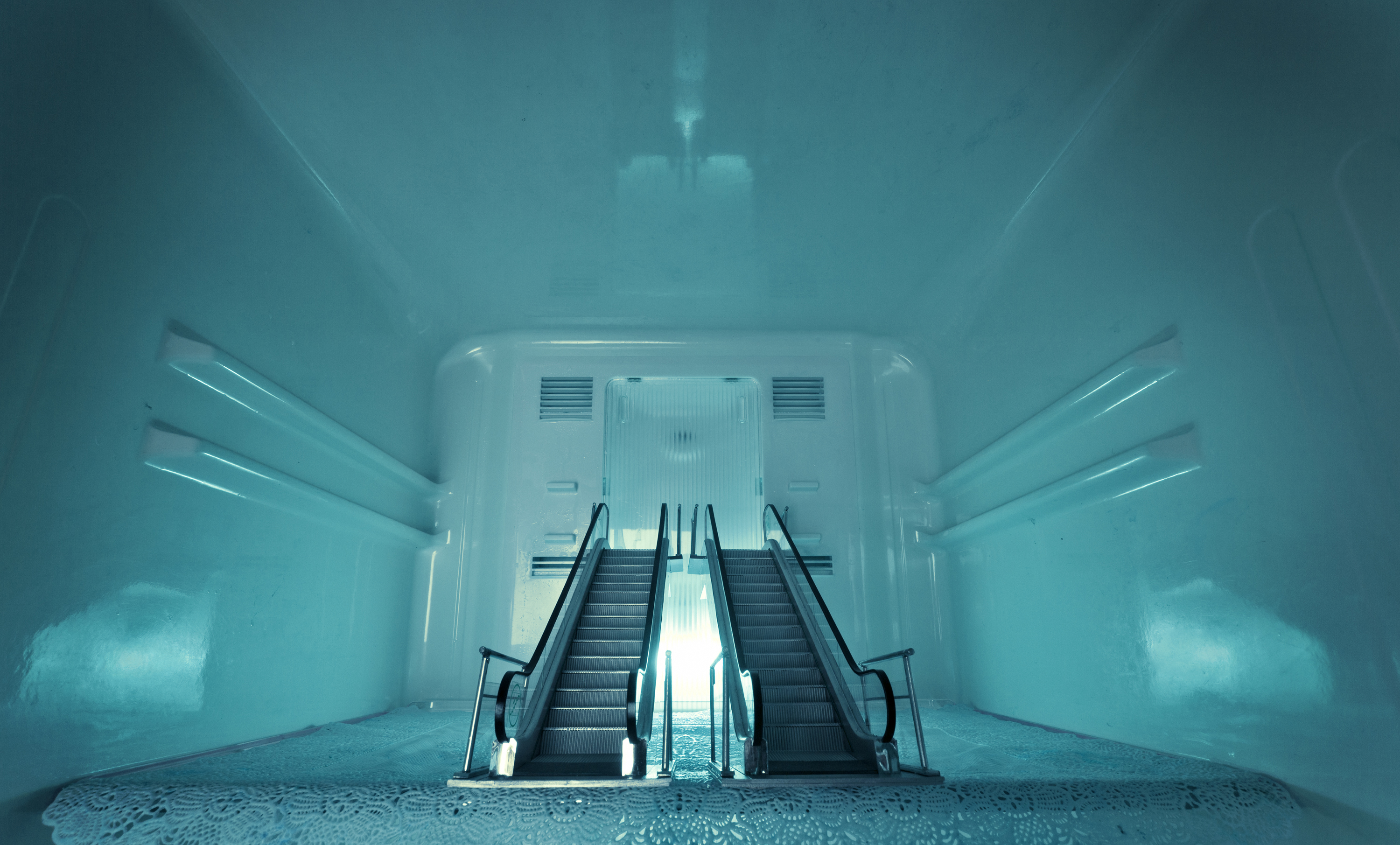 A surreal scene of two escalators emerging from the floor of an empty, turquoise-painted room, leading upwards. The lighting gives the room an ethereal, futuristic feel.