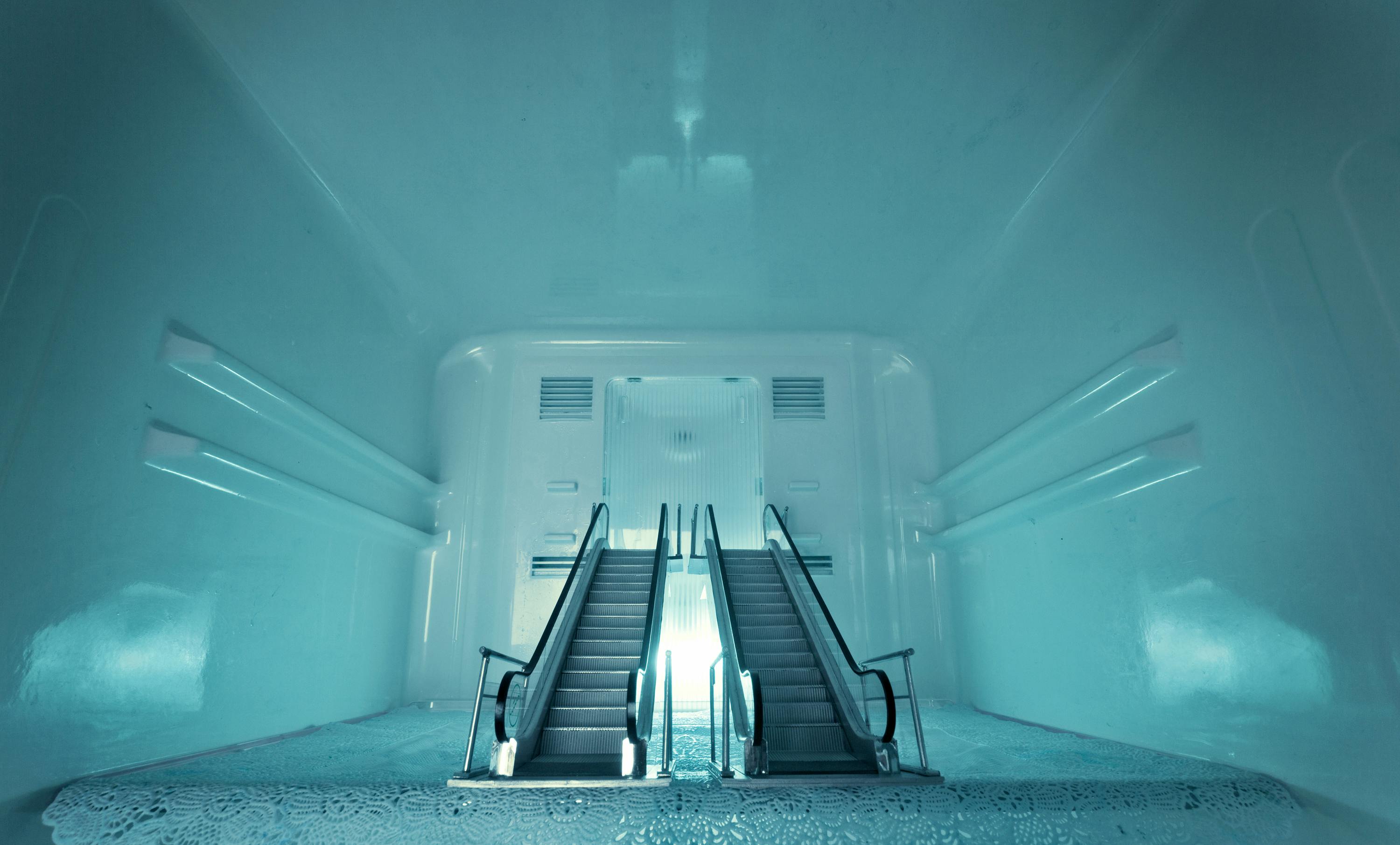 A surreal scene of two escalators emerging from the floor of an empty, turquoise-painted room, leading upwards. The lighting gives the room an ethereal, futuristic feel.