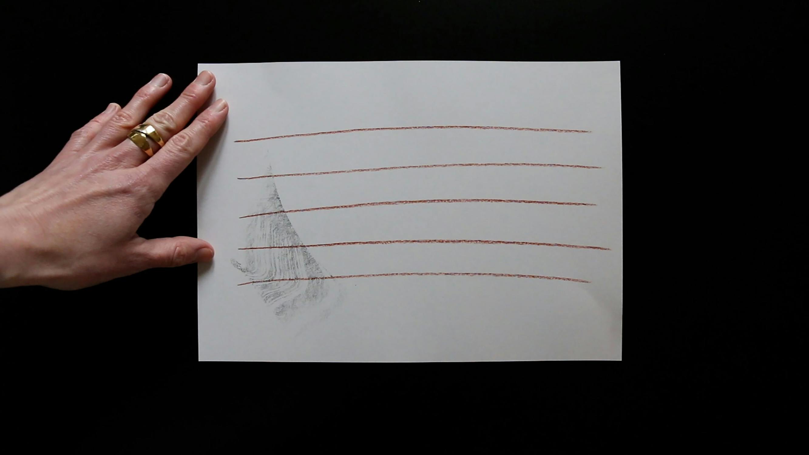 A hand holds a sheet of paper with six horizontal red lines and a shaded triangular area on a black background. The person wears a wedding band and ring on the left hand.