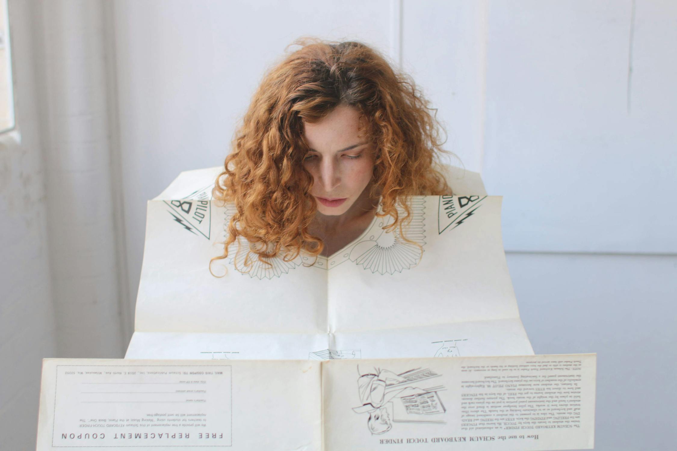 A person with curly red hair is looking down at a large sheet of paper, possibly a map or a newspaper, in a bright room. The paper features illustrations and text.