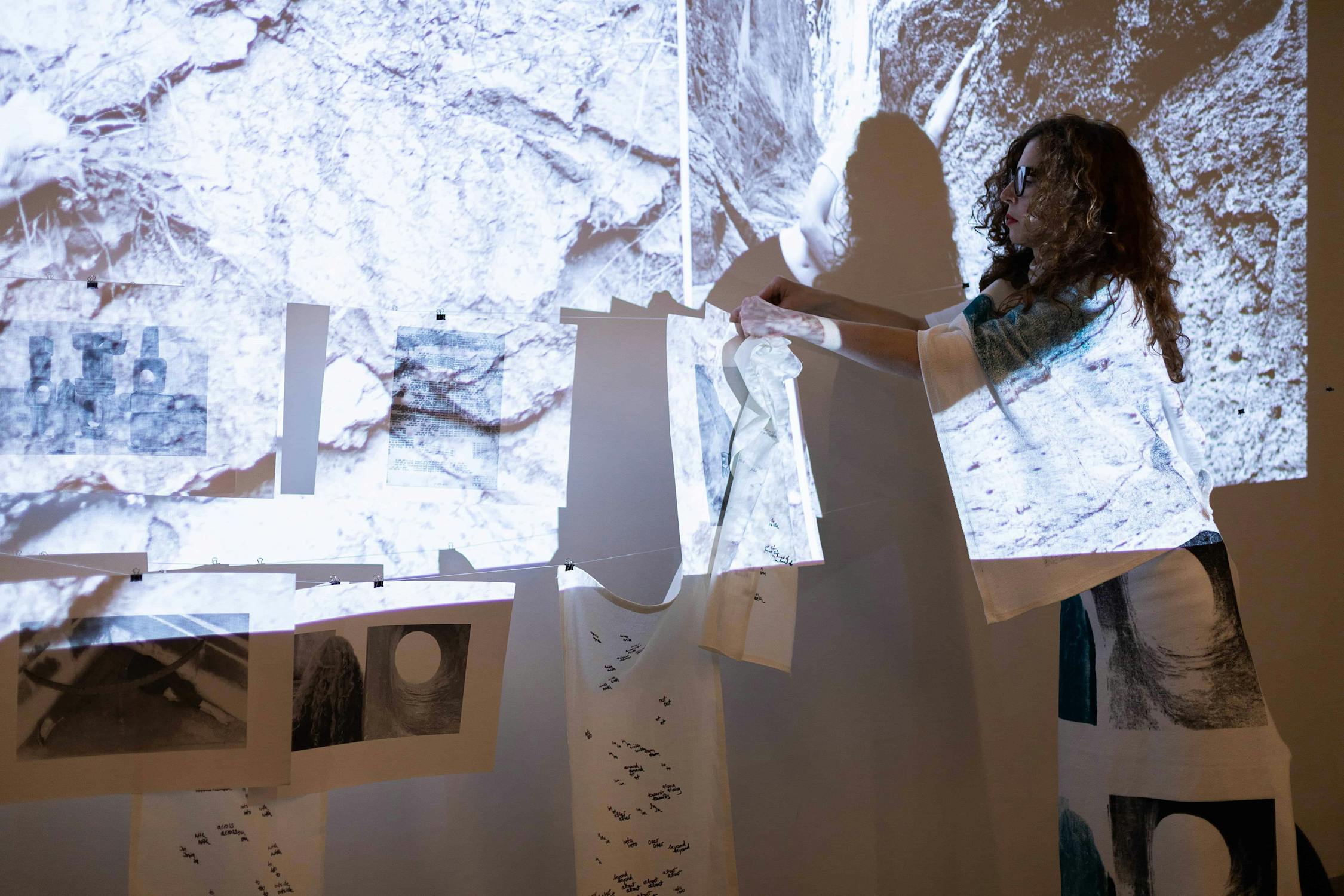 A person interacts with a wall display of projected images and hanging papers. The scene has an artistic and creative setting, with abstract imagery and a dimly lit atmosphere.