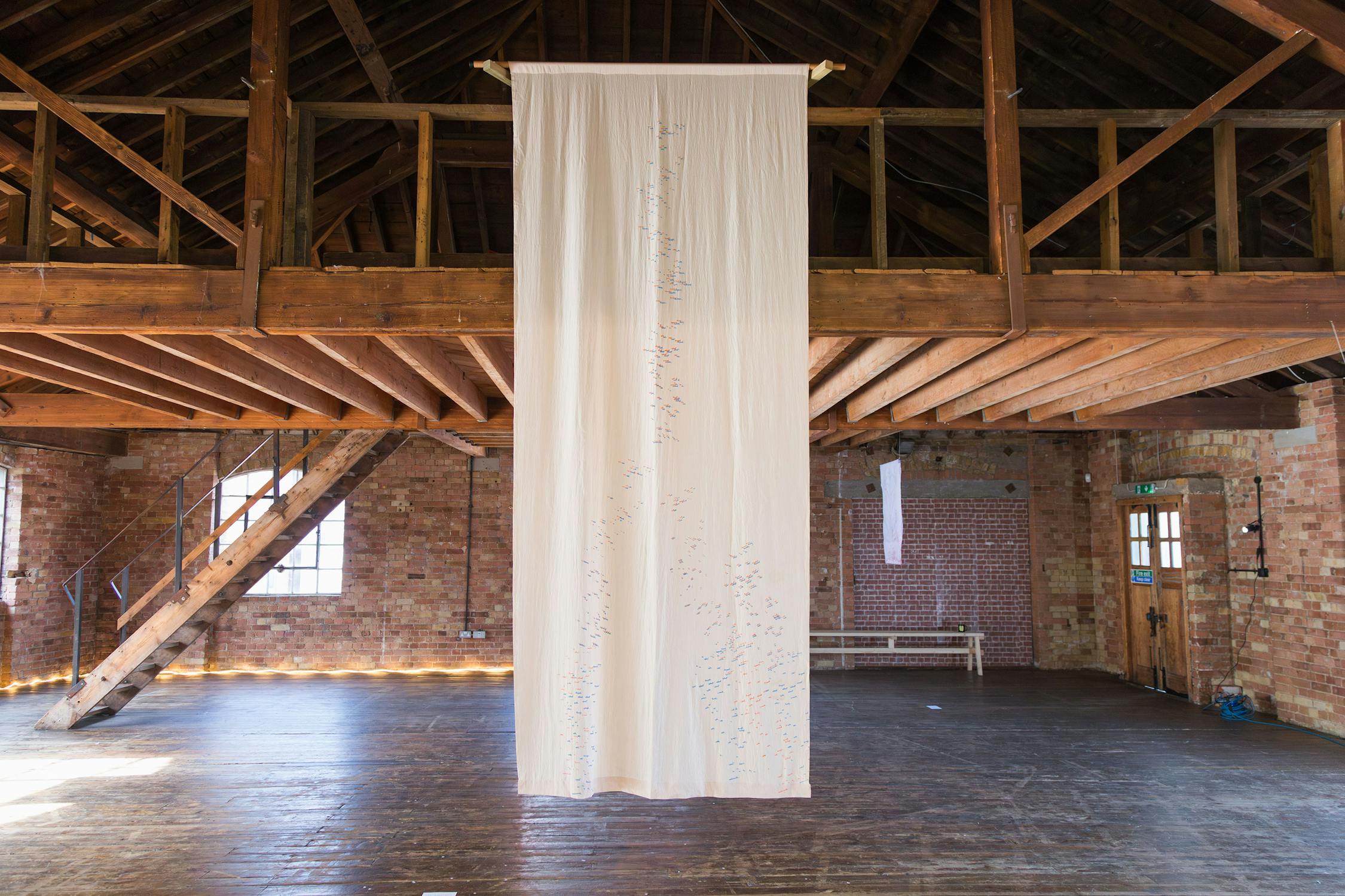 A long, white fabric banner with small, colourful details hangs from a wooden beam in a rustic, open space with exposed brick walls and wooden floors. Sunlight filters through a window, creating a serene atmosphere.