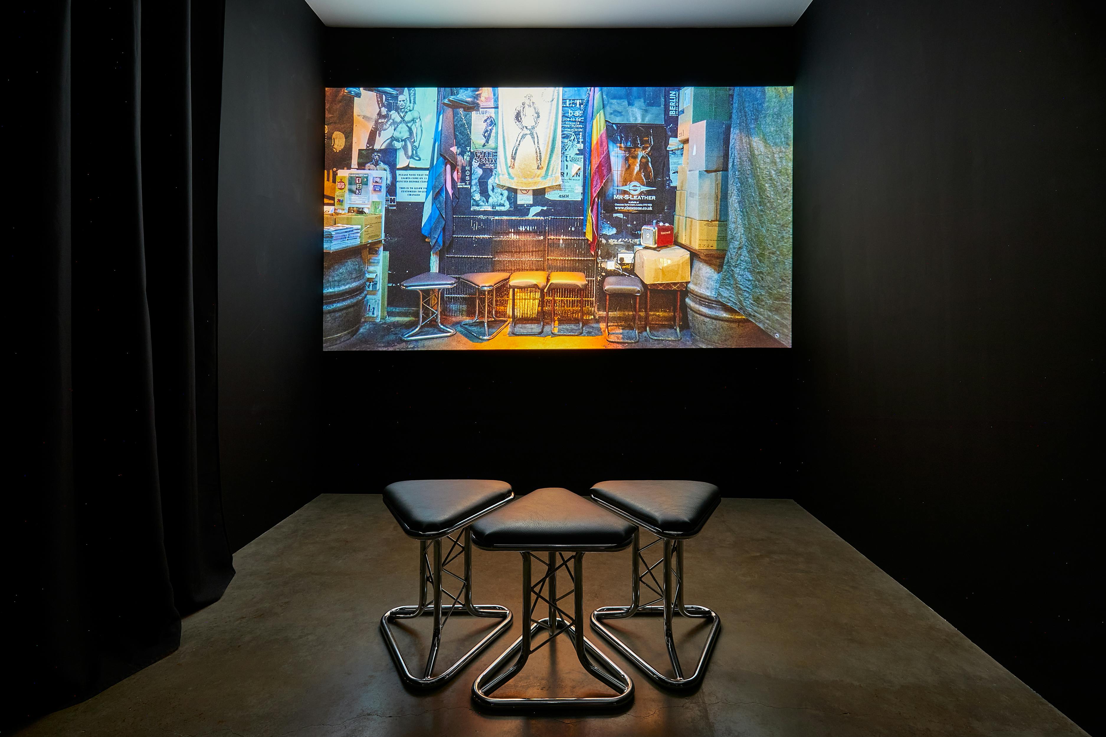 A dark room features three triangular stools with metal legs facing a projected image on the wall. The image depicts a dimly lit bar or pub with chairs, tables, and various decorations.