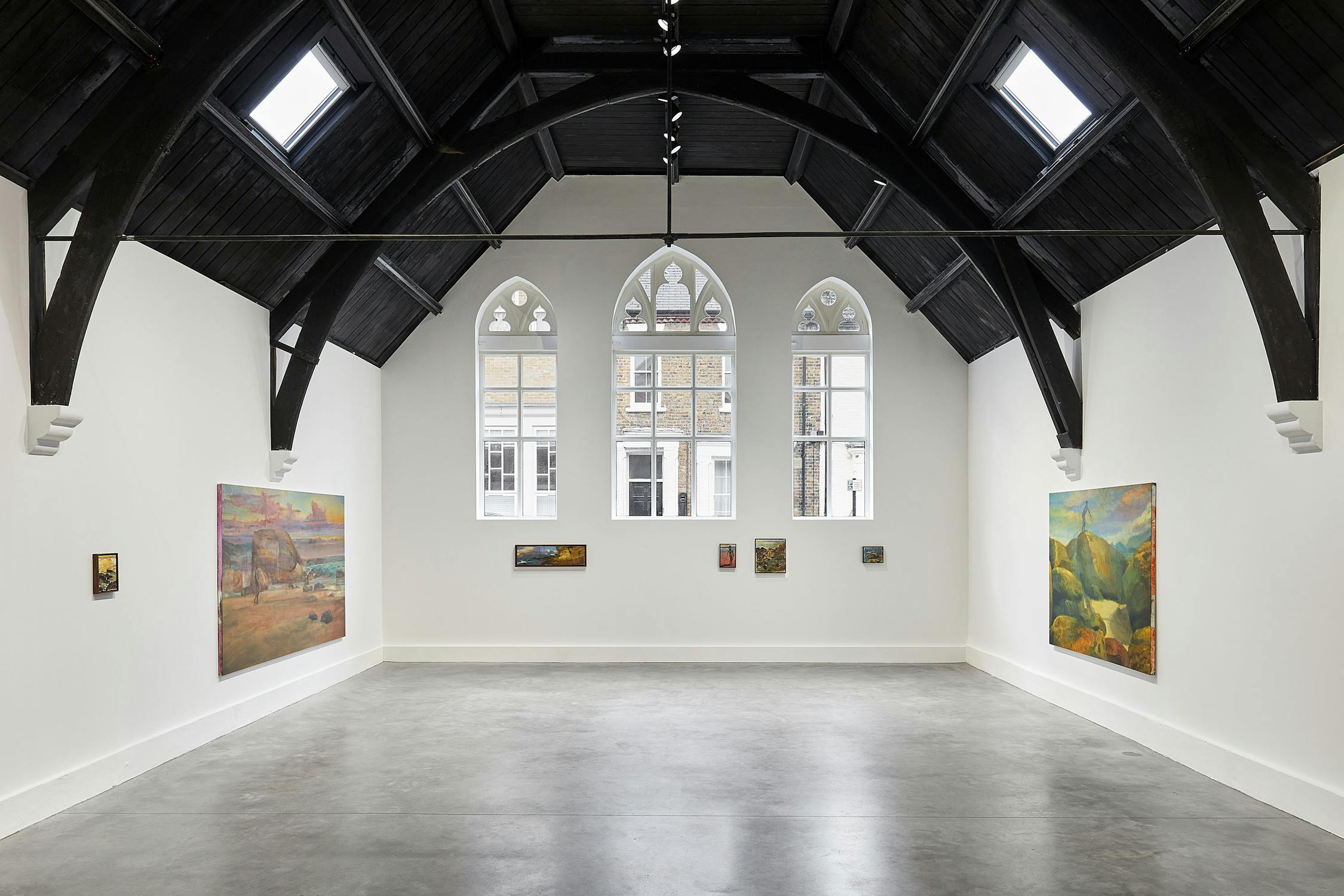 A spacious art gallery with white walls and a high, dark wooden vaulted ceiling. Several colourful paintings are displayed on the walls. Large arched windows at the far end let in natural light, illuminating the polished concrete floor.