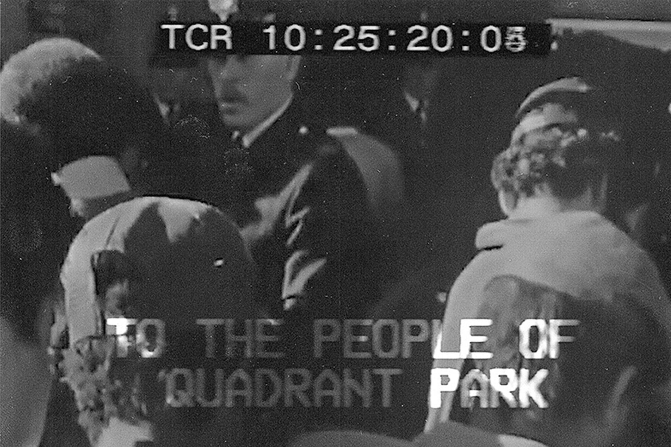 A grainy, black-and-white photo shows several people standing close together. A police officer is partially visible among them. Text overlaid on the image reads, "TO THE PEOPLE OF QUADRANT PARK."