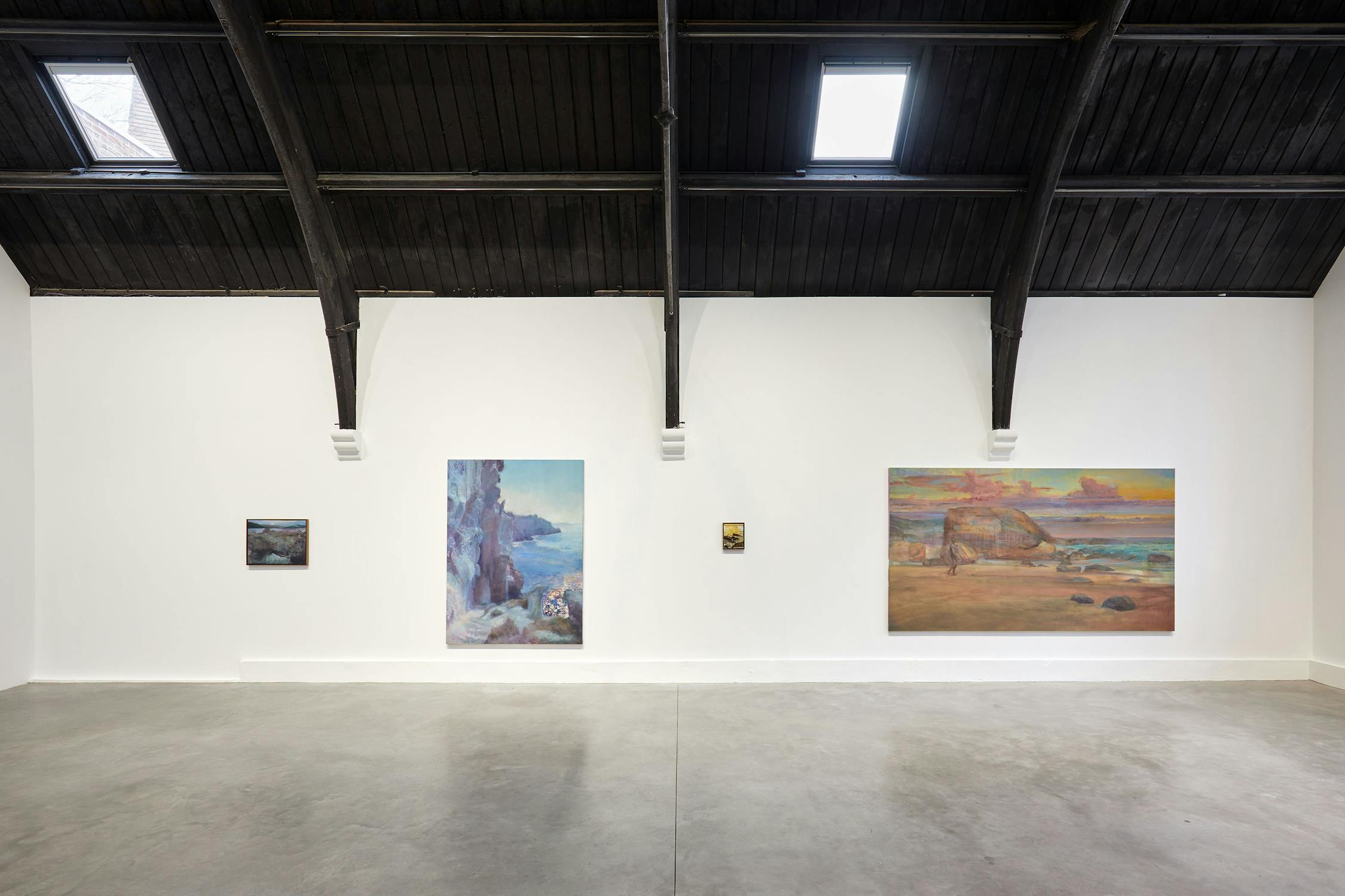 A gallery room with a high, dark wooden ceiling displays five paintings by Jake Grewal on a white wall. The artworks vary in size and depict landscapes and nature scenes, including cliffs and a beach at sunset. The floor is smooth concrete.